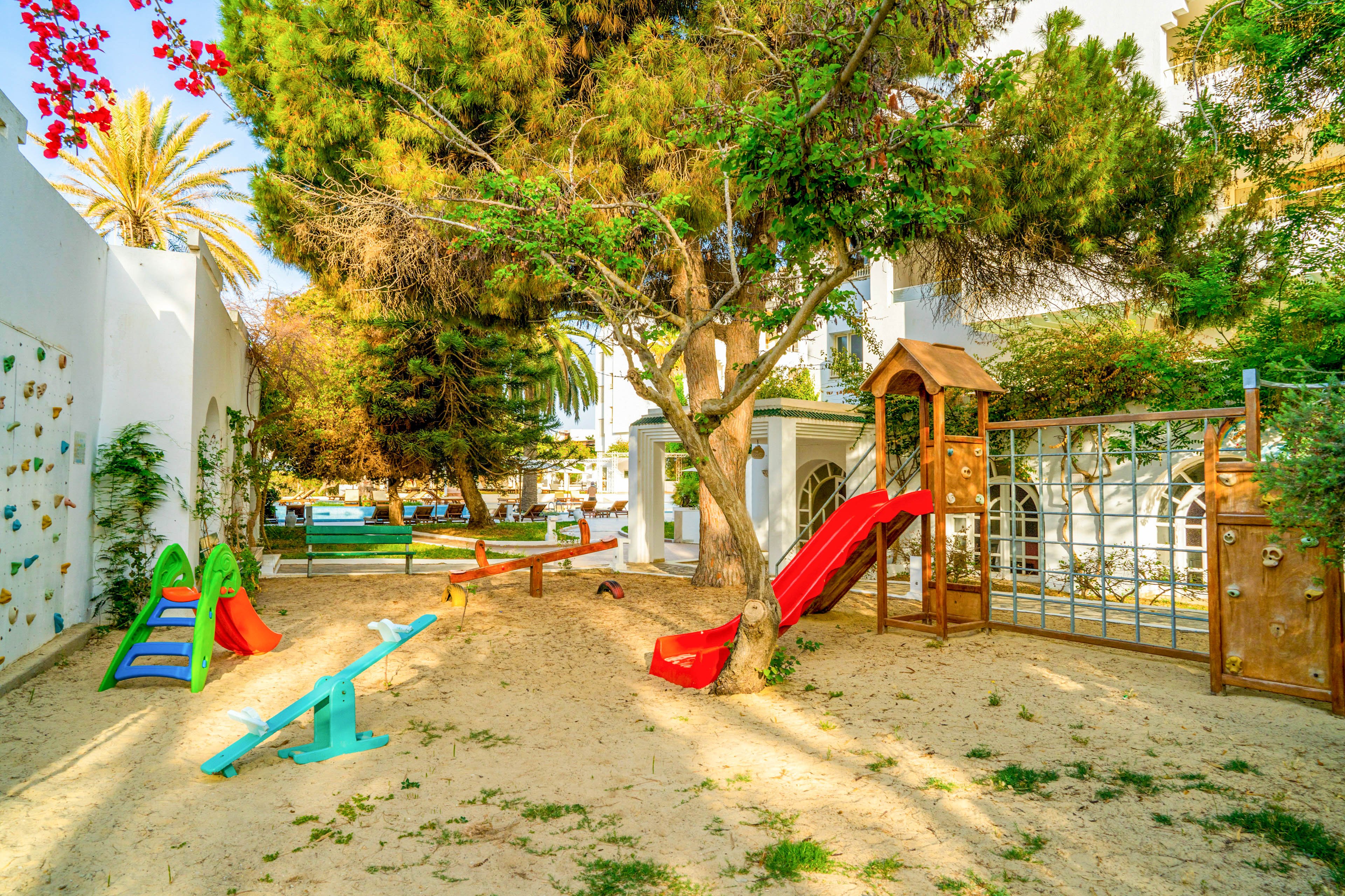 A bright outdoor playground is set in a sandy yard surrounded by trees and white walls. Colorful play structures, including a red slide, seesaw, and small climbing frames, are arranged under the shade of tall pines. Sunlight filters through the foliage, casting dappled shadows across the sand. The scene feels warm and inviting, ideal for children’s play in a residential or school setting.