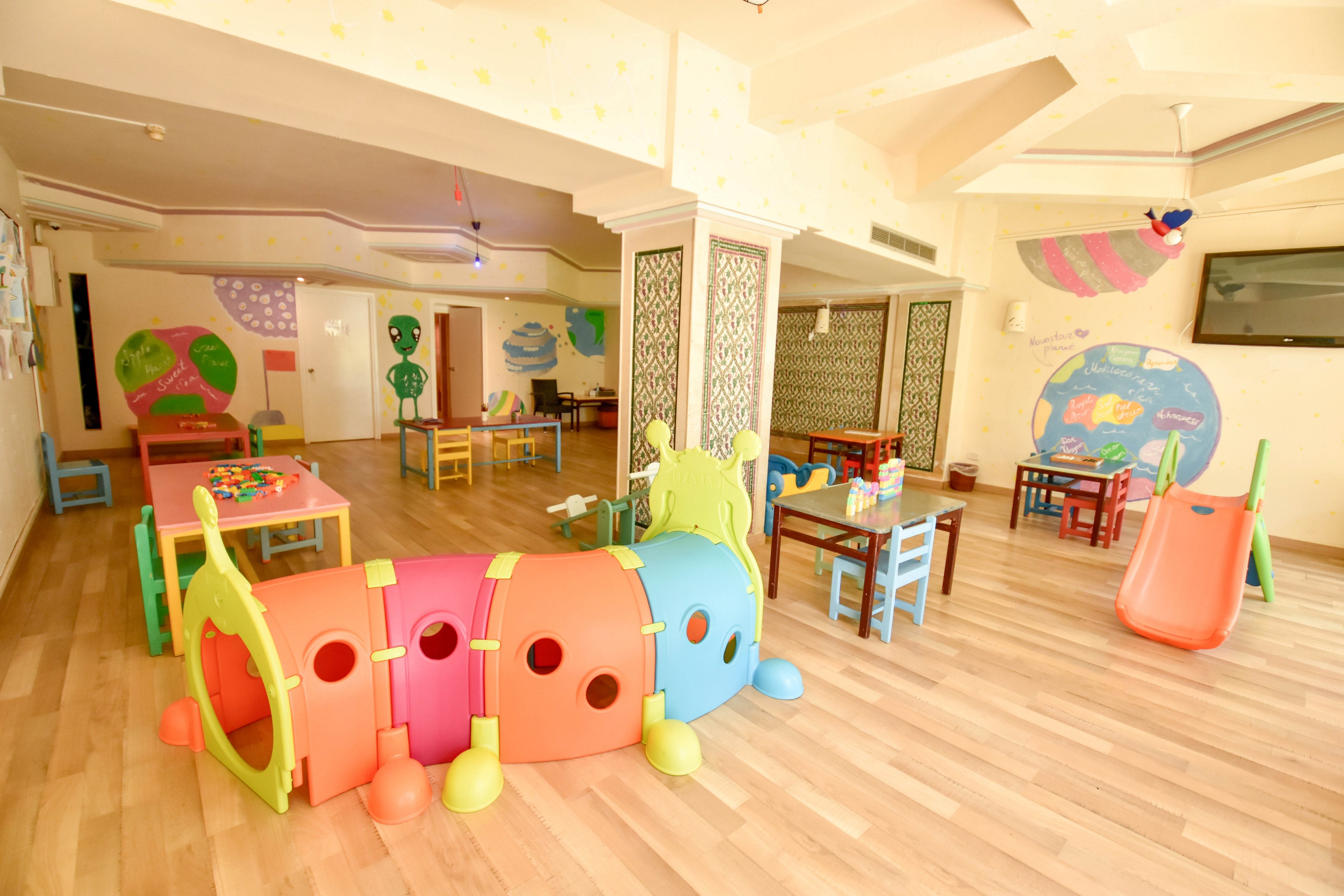 A colorful indoor preschool playroom features a large caterpillar tunnel structure in the foreground. Small tables and chairs are arranged around the room for children’s activities and crafts. A plastic slide, toy storage, and various play items are scattered throughout the space. The walls are decorated with playful murals and shapes, creating a cheerful and inviting environment.