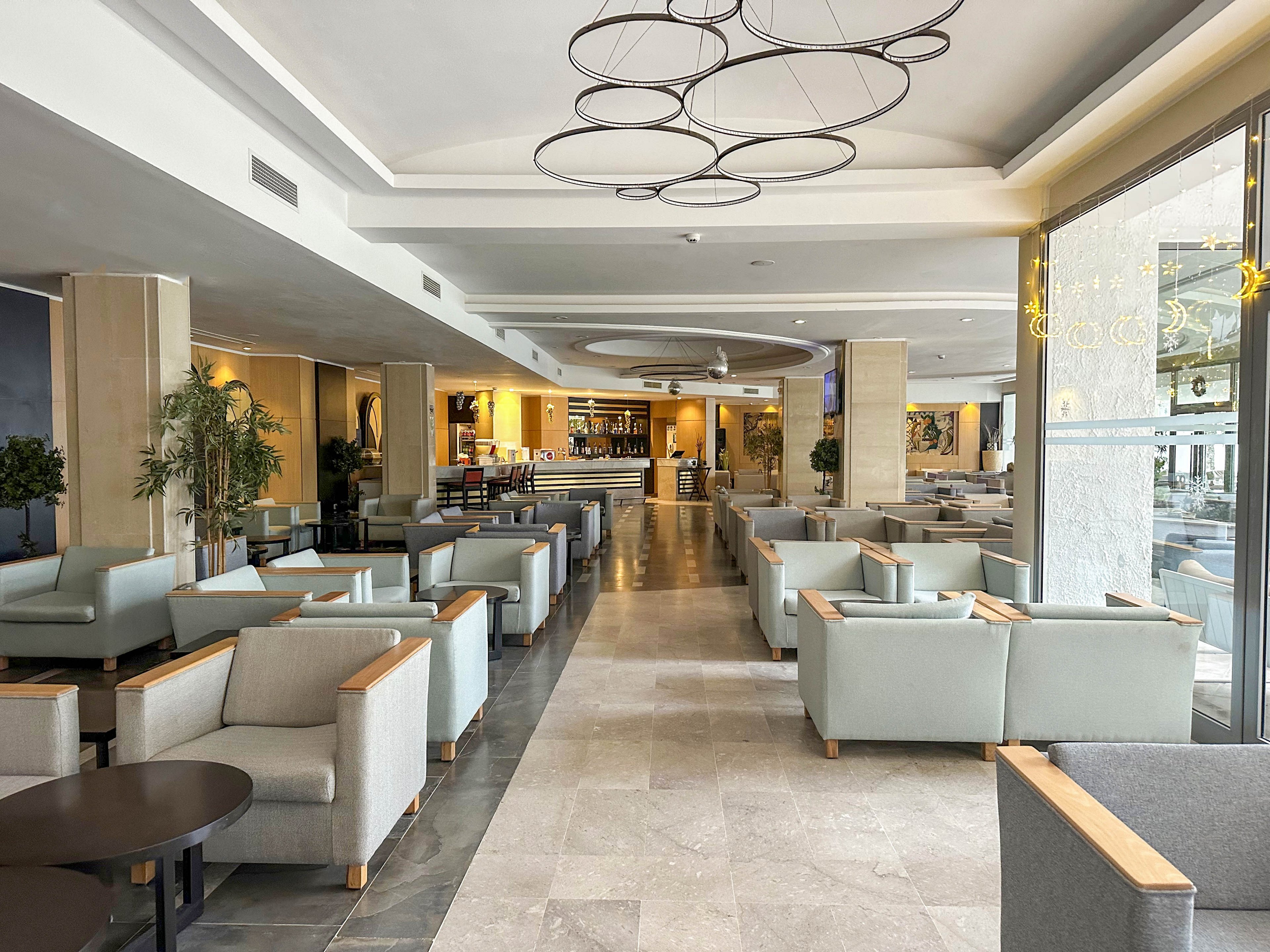 A spacious modern hotel lobby lounge features rows of light gray armchairs and low coffee tables arranged symmetrically. The setting includes a central aisle leading toward a well-lit bar area at the back of the room. Large windows on the right allow natural light to brighten the neutral-toned interior, complemented by circular ceiling light fixtures. Potted plants and warm accent lighting add a welcoming, contemporary atmosphere.