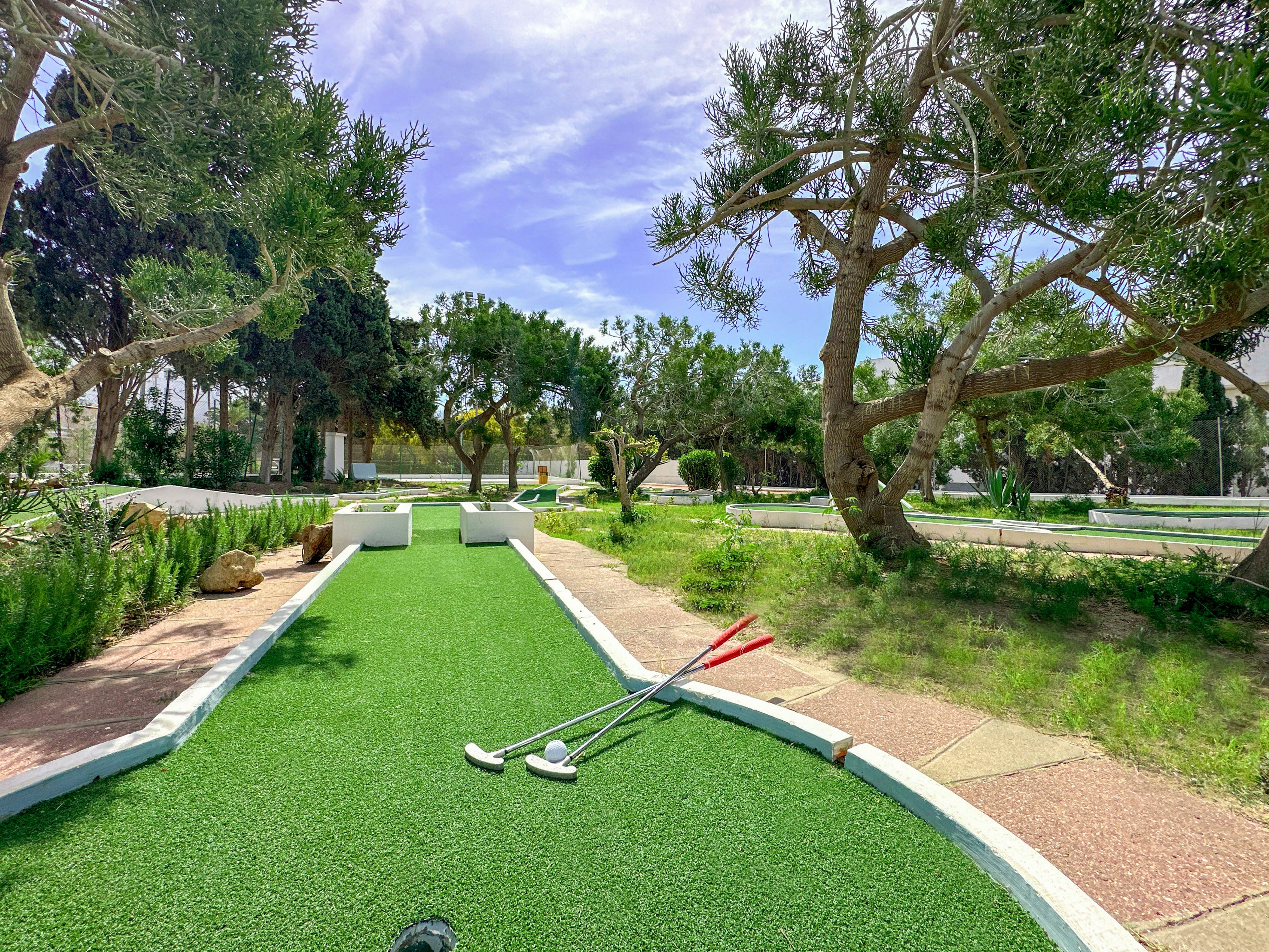 An outdoor mini golf course is shown under a bright sky, with a green artificial turf lane leading toward a simple white obstacle. Several putters and a golf ball rest on the turf in the foreground. The course is surrounded by trees, shrubs, and a mix of paved and grassy areas. The scene feels calm and leisurely, suggesting a casual recreational setting.