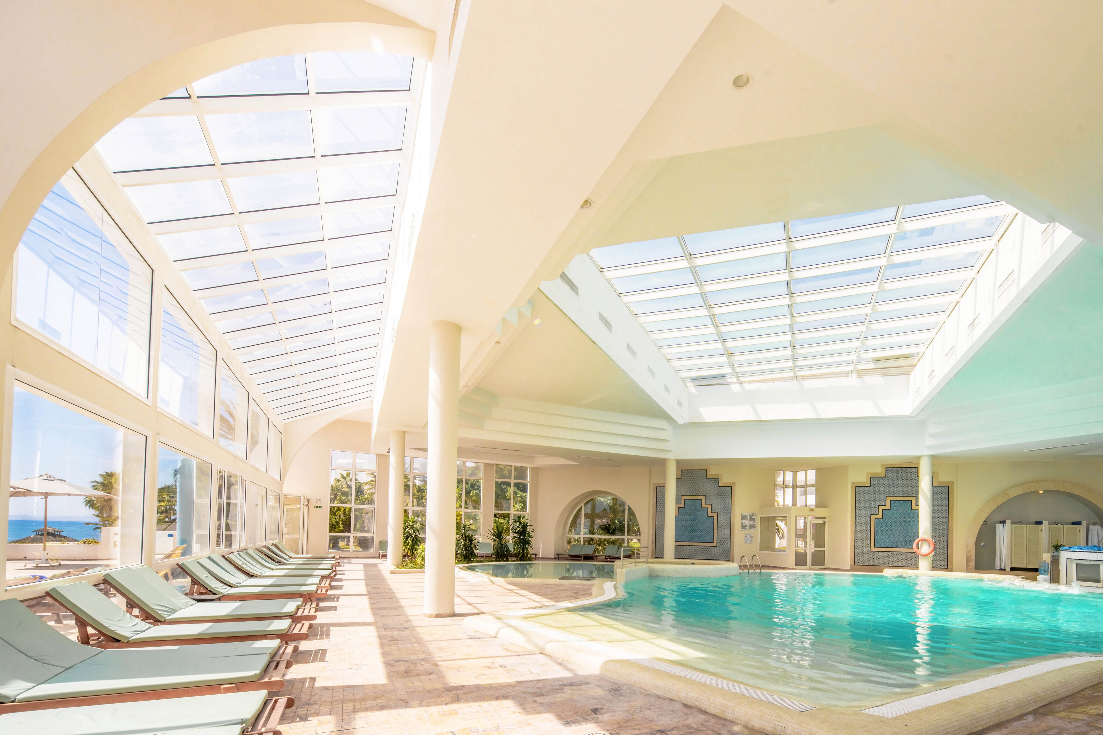 A spacious indoor swimming pool is bathed in natural light from large skylights and floor-to-ceiling windows. Multiple lounge chairs line the tiled deck, creating a relaxing resort atmosphere. The architecture features high arches, white columns, and soft pastel accents around the pool area. Outside the windows, a glimpse of blue sky and water suggests a coastal or beachfront location.