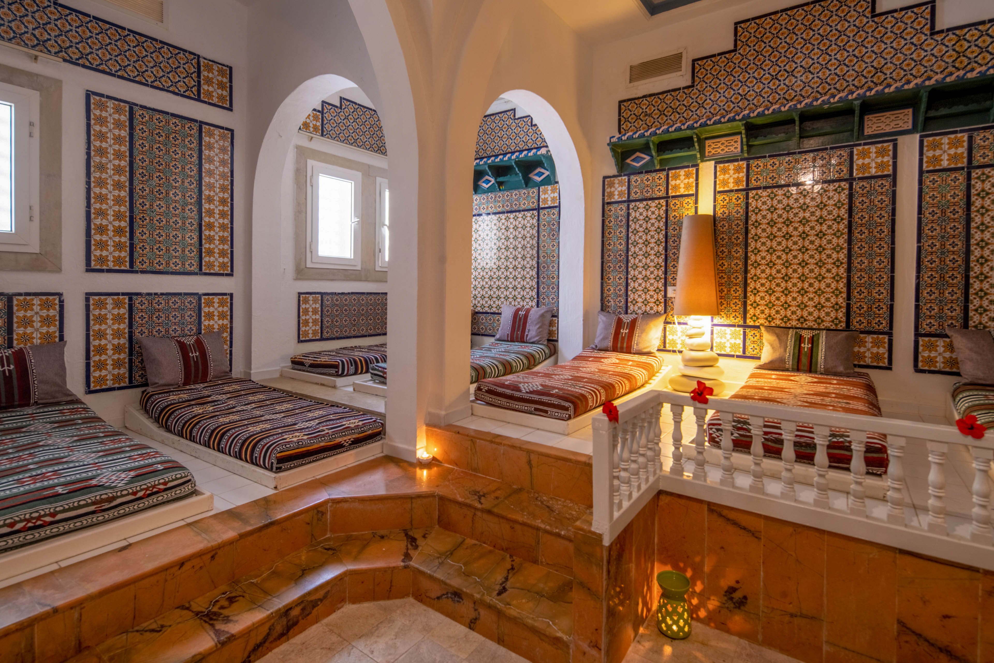 A warm, inviting lounge features multiple low beds lined with striped cushions and textiles. The walls are decorated with intricate geometric tiles in rich colors, framed by white arches and windows. Soft ambient lighting, including candles and a lantern, creates a relaxing atmosphere around the central tiled platform. The overall scene evokes a traditional, cozy interior ideal for rest and social gatherings.