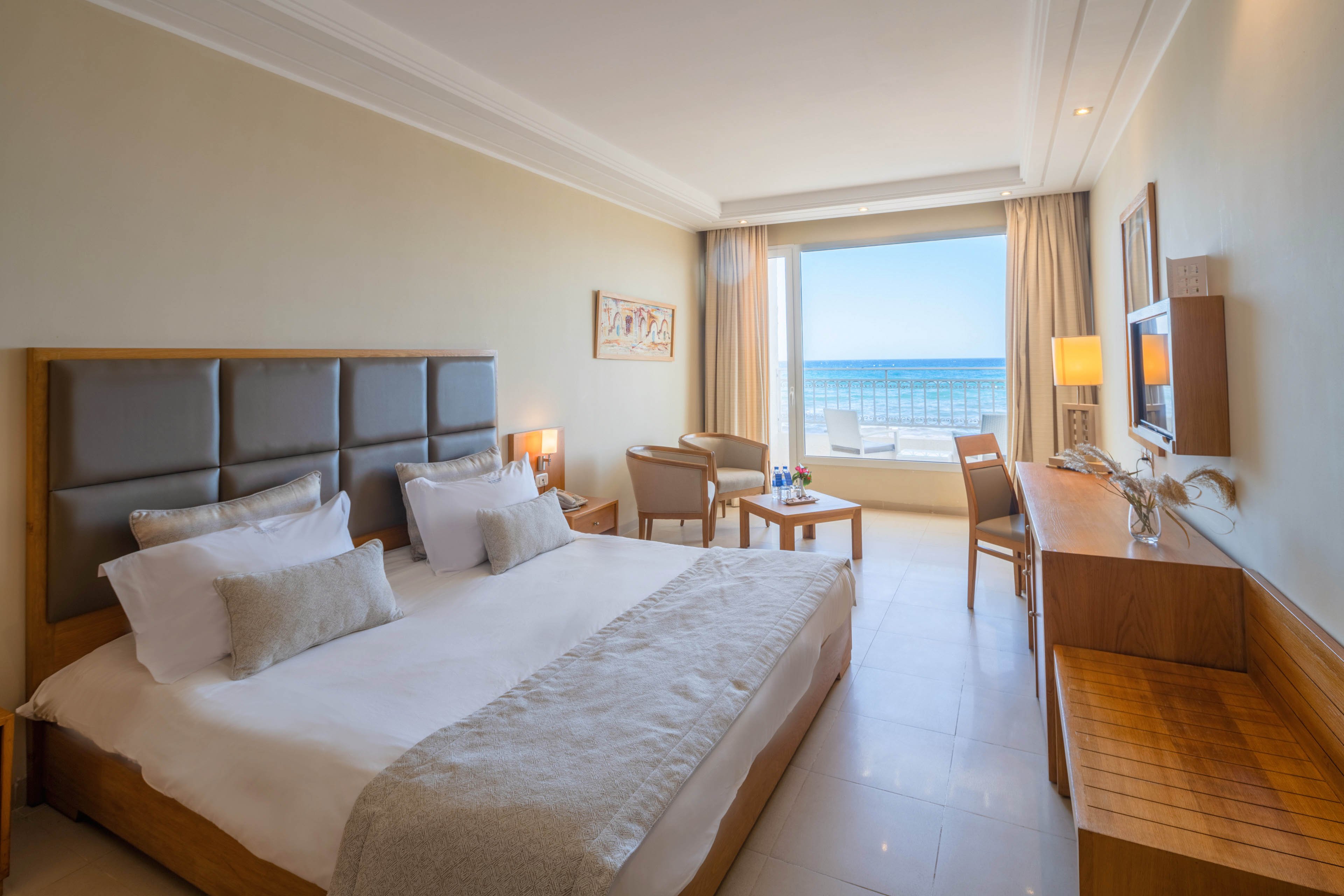 A modern hotel room features a large neatly made bed with neutral-toned linens and cushions. The space includes a wooden desk, chair, and small seating area with two armchairs and a round table. Large windows with light curtains open to a balcony overlooking a bright blue ocean and clear sky. Warm lighting, soft colors, and minimal decor create a calm, inviting atmosphere ideal for relaxation.