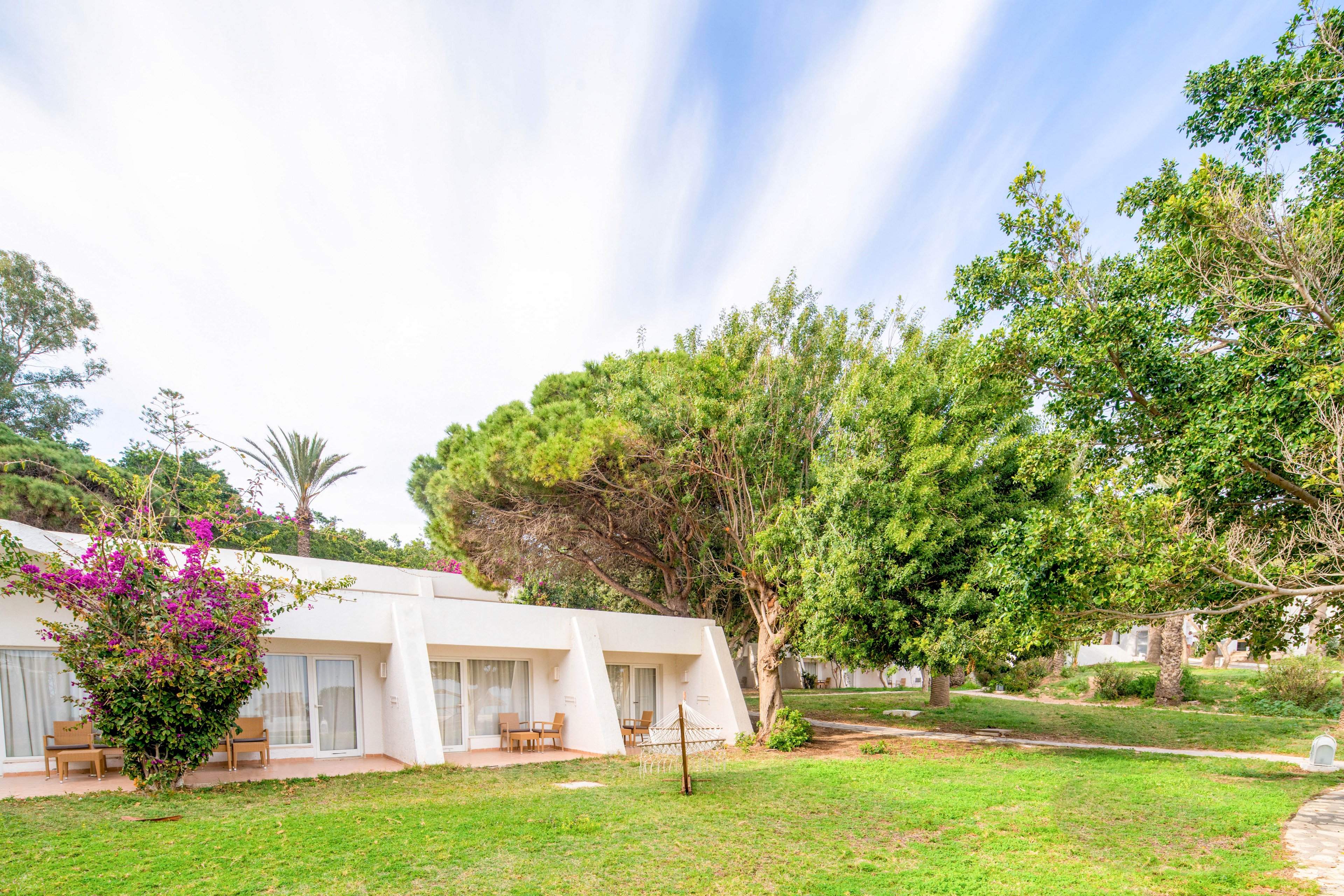 Single-story white bungalows sit in a green garden setting under a bright sky. Large trees and flowering shrubs surround the buildings, creating a tranquil, natural atmosphere. Patio chairs and glass doors suggest private outdoor seating areas for guests. The scene feels peaceful and inviting, ideal for a vacation or retreat.