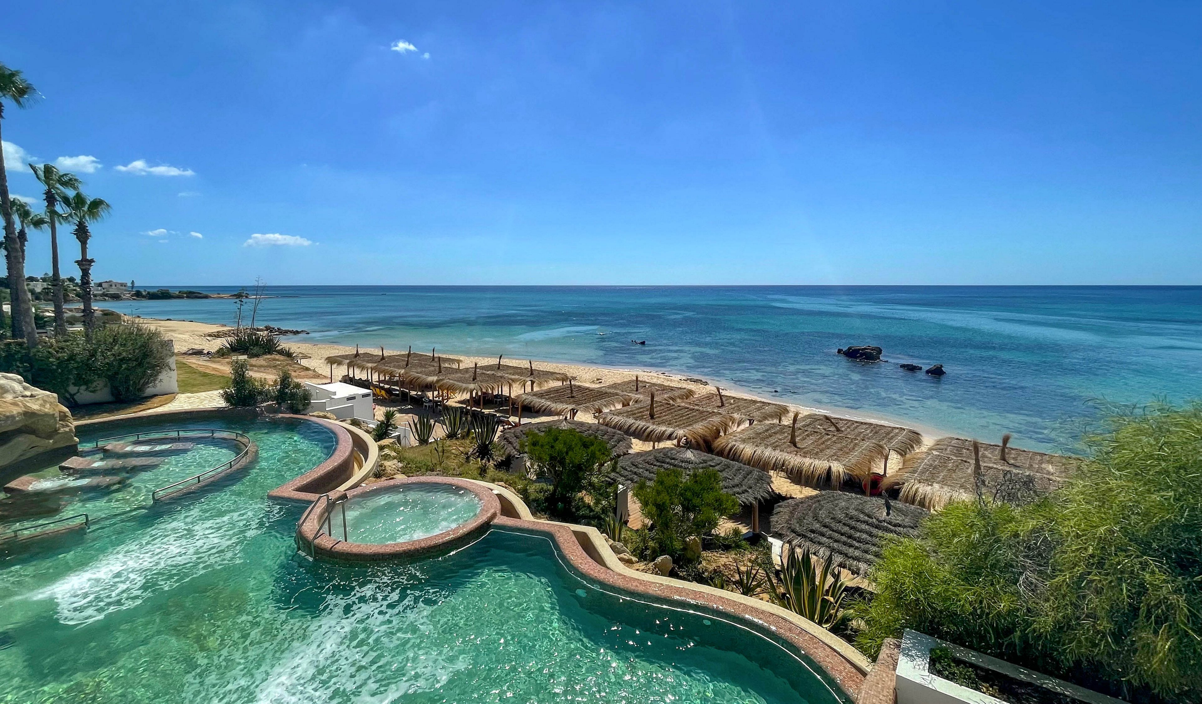 A series of turquoise infinity pools curves along the edge of a coastal resort. Thatched cabanas and wooden structures line the sandy beach below, leading toward the calm blue ocean. Palm trees and lush greenery frame the scene under a clear, sunny sky. The tranquil setting suggests a luxurious, relaxing seaside getaway.