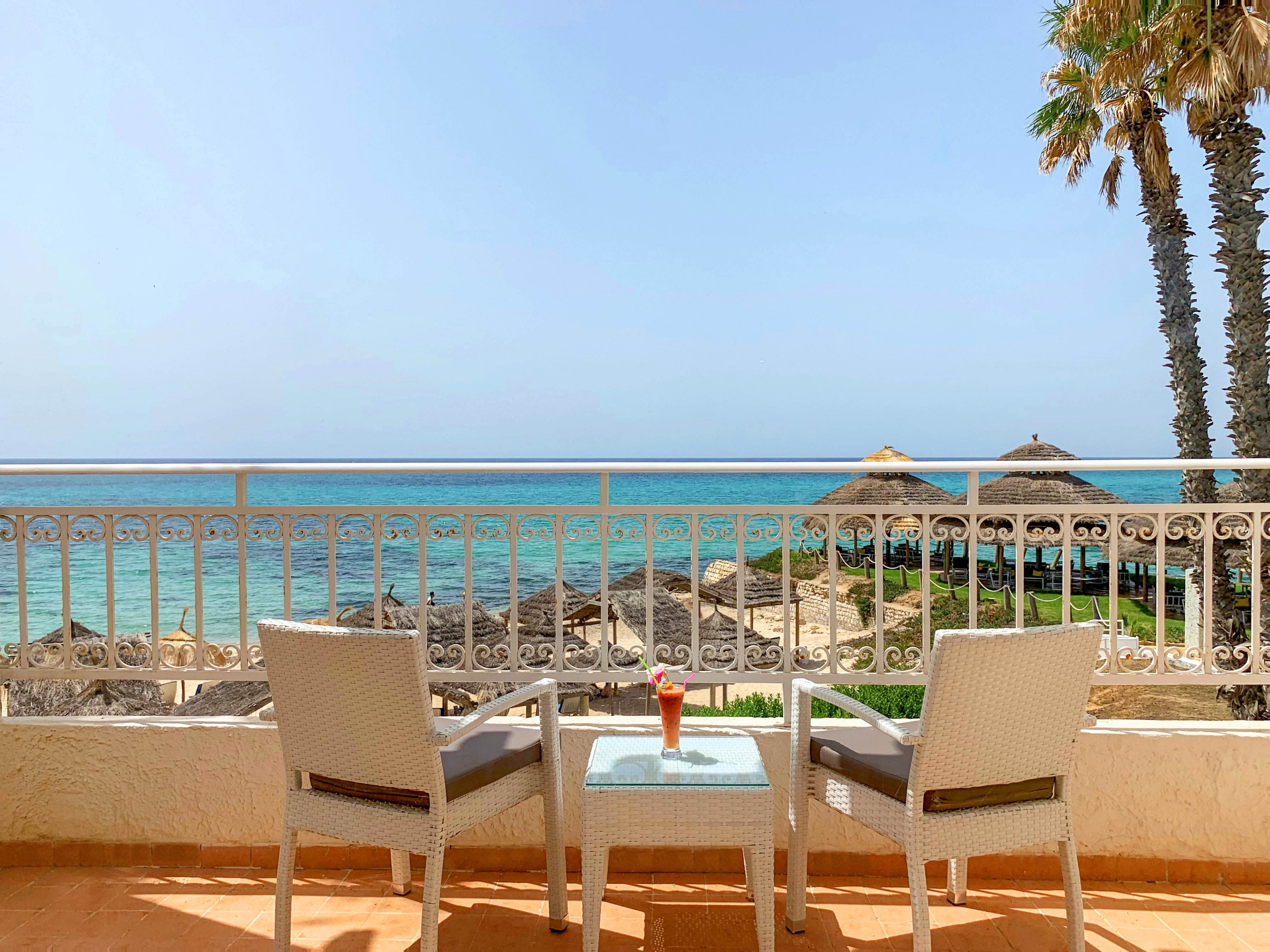 A bright seaside terrace overlooks a calm blue ocean. Two white chairs face a small glass-top table holding a single colorful drink. Beyond the railing, thatched umbrellas and palm trees line the sandy shoreline. The clear sky and warm light create a relaxed, vacation atmosphere.