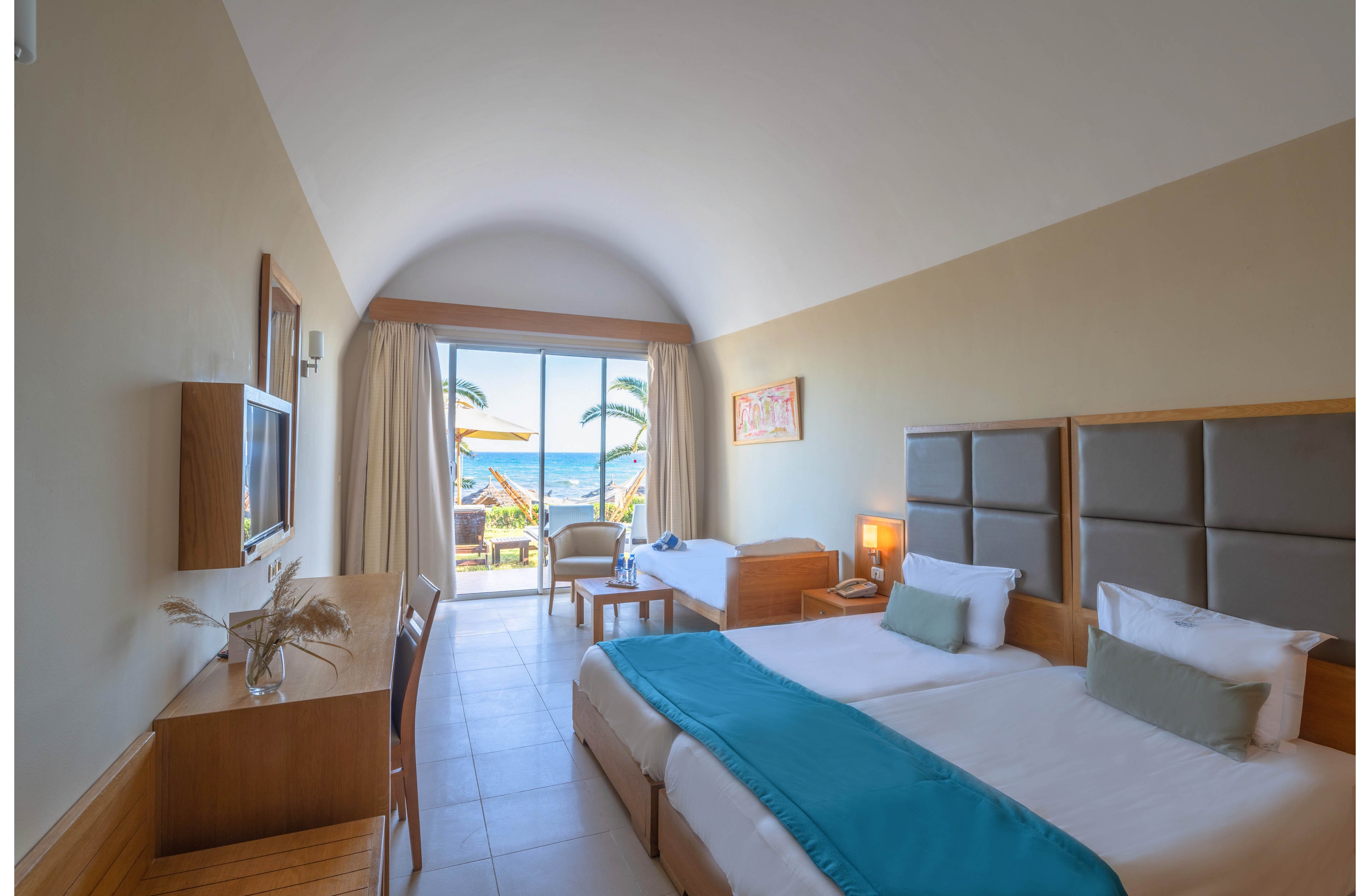 A modern hotel room features two neatly made beds with teal runners and padded headboards. The space includes a wooden desk, chair, and a small sitting area with a table and additional bed near the window. Large glass doors open to a balcony overlooking palm trees and the ocean, allowing natural light to fill the room. Soft neutral walls and light tile flooring create an airy, relaxing atmosphere.