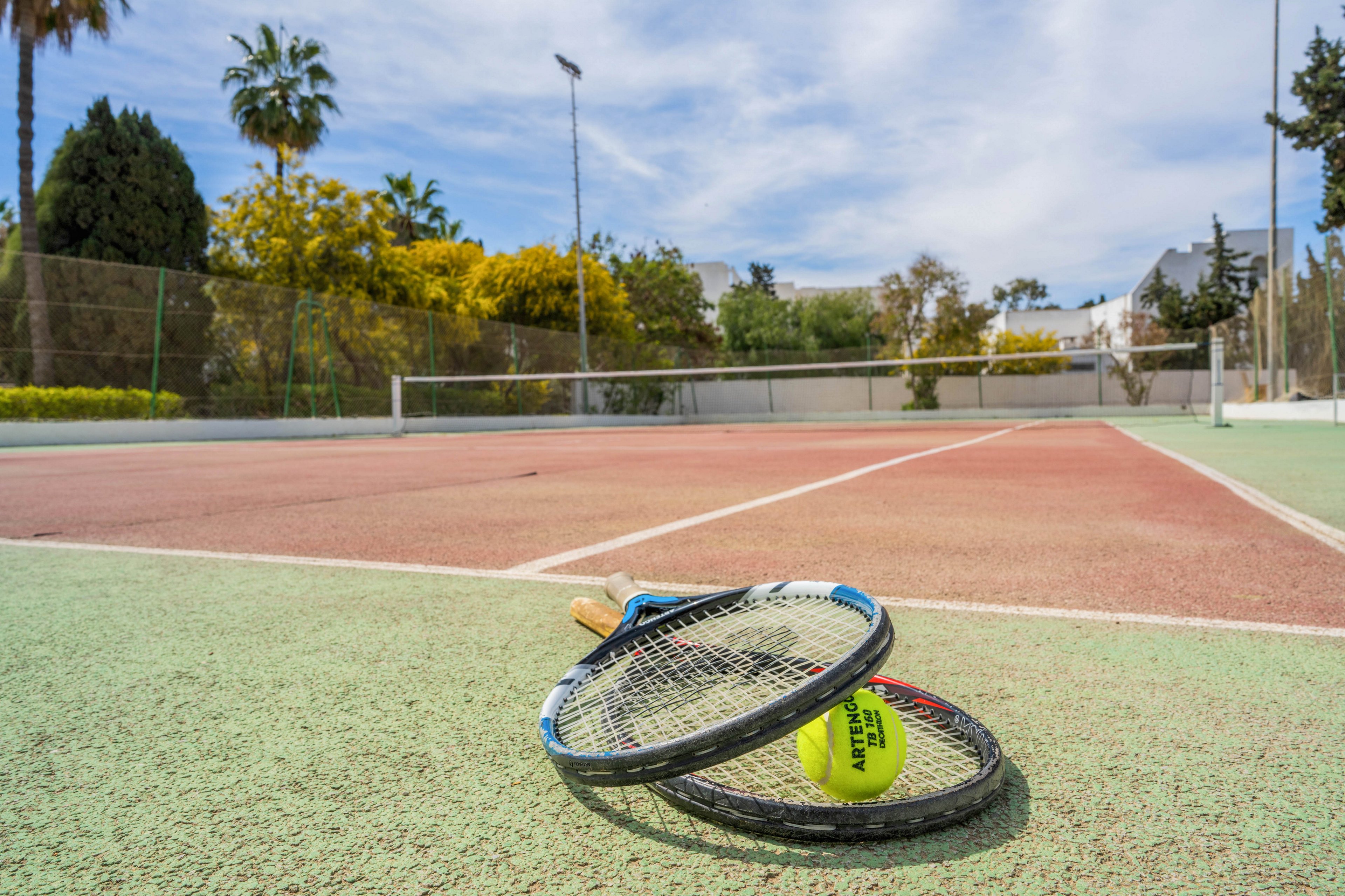 Two tennis rackets and a tennis ball lie on the surface of an outdoor court. The scene is set on a bright day with a clear sky and scattered clouds. A net stretches across the court, and trees and palm trees surround the area. The image emphasizes leisure sports and a relaxed, sunny atmosphere.