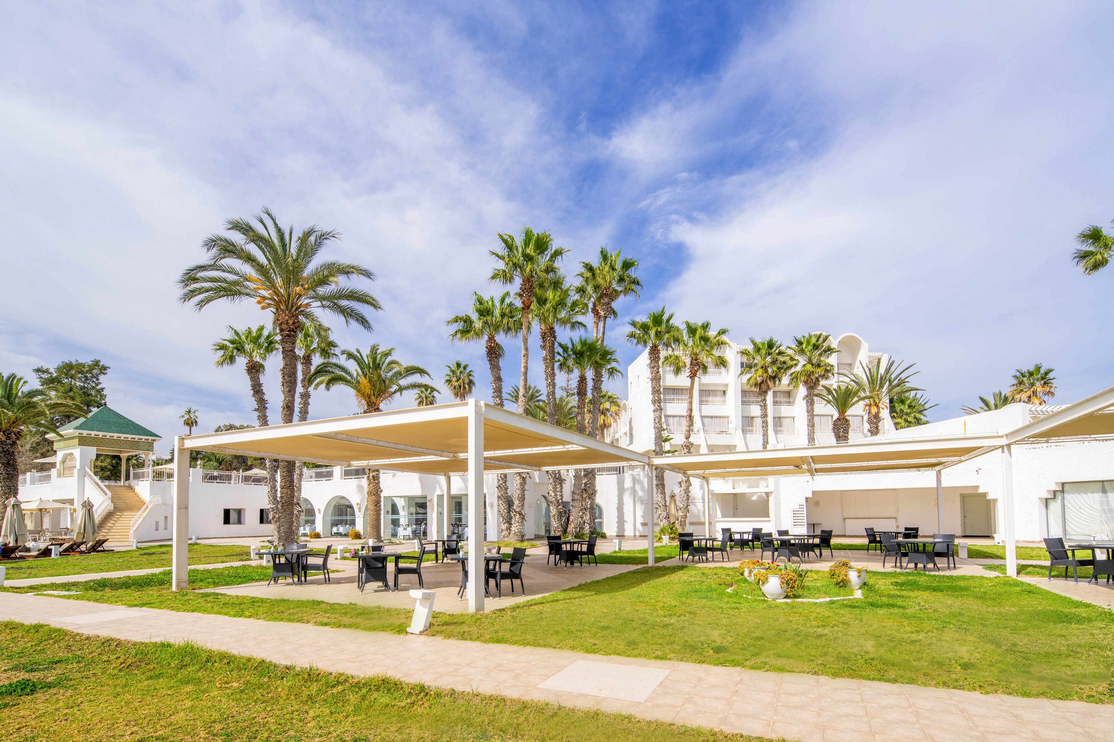 An outdoor courtyard of a seaside resort features open pergolas with tables and chairs arranged on a manicured lawn. Tall palm trees rise behind the seating area, framing the white hotel buildings in the background. The sky is bright with scattered clouds, creating a warm and inviting atmosphere. Pathways and trimmed grass add to the clean, organized look of the leisure space.
