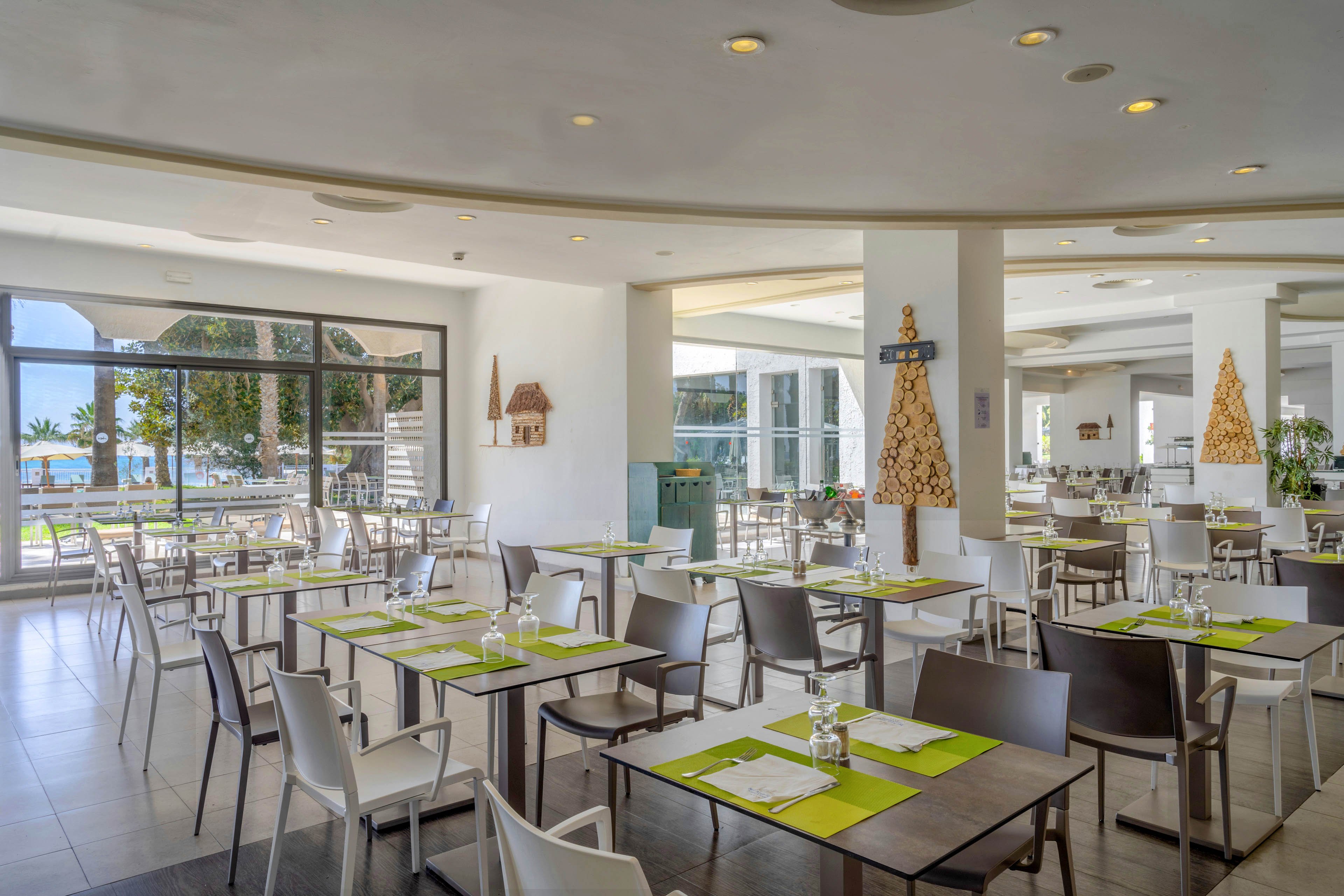 A modern restaurant dining area is set with neatly arranged tables and chairs, each table topped with green placemats and glassware. Large windows allow natural light to flood the space and reveal a view of the sea and palm trees outside. Minimalist holiday decorations, including cone-shaped and tree-shaped ornaments, add a festive touch to the white interior. The overall atmosphere is clean, airy, and inviting for guests.