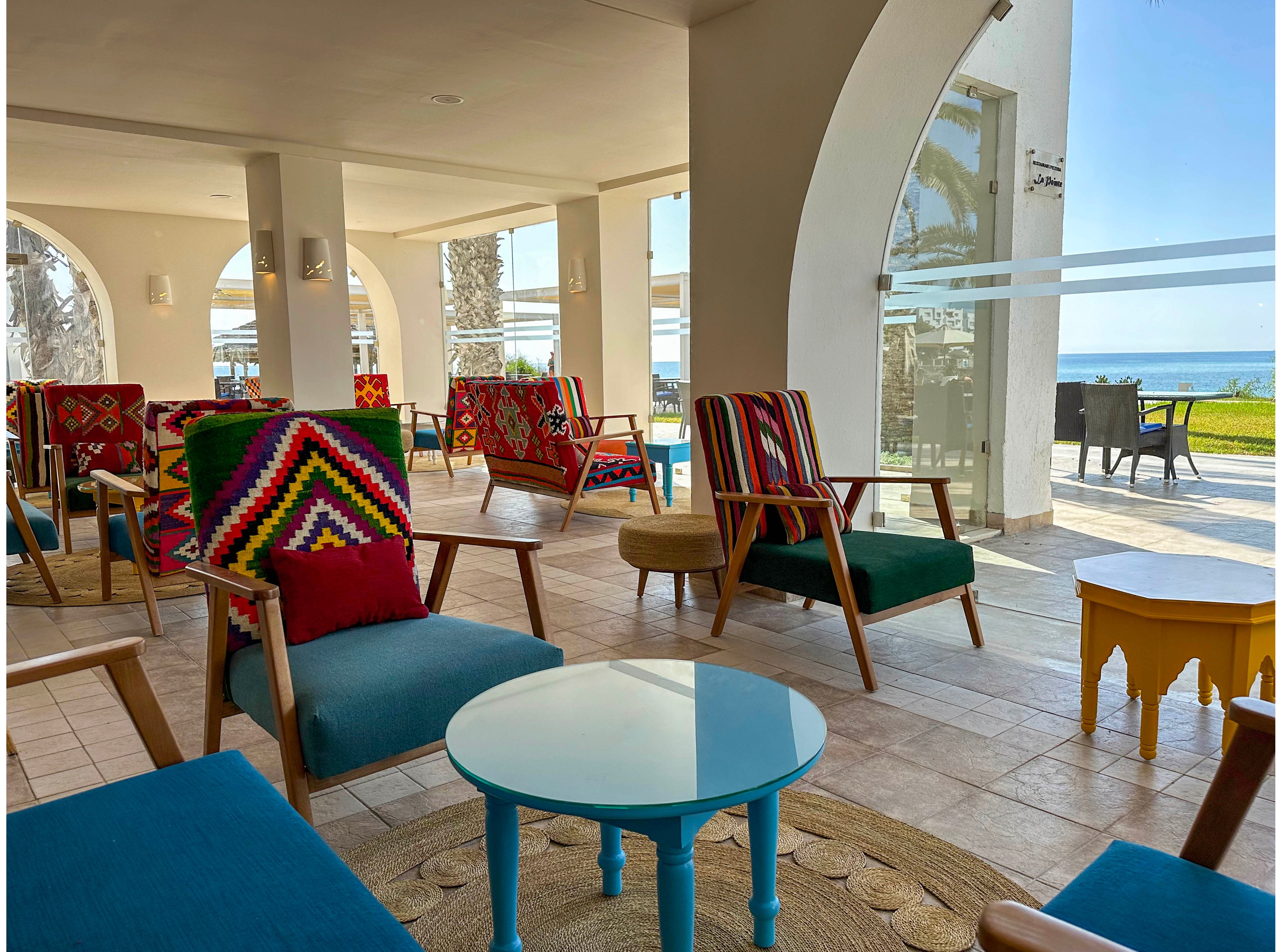 A bright indoor lounge area features modern wooden armchairs with teal cushions and vibrant geometric-patterned backs. A round turquoise coffee table sits on a woven rug, surrounded by additional seating and small side tables. Large arched openings reveal palm trees, a green lawn, and the sea in the background. Natural light floods the tiled space, creating a relaxed, coastal atmosphere.