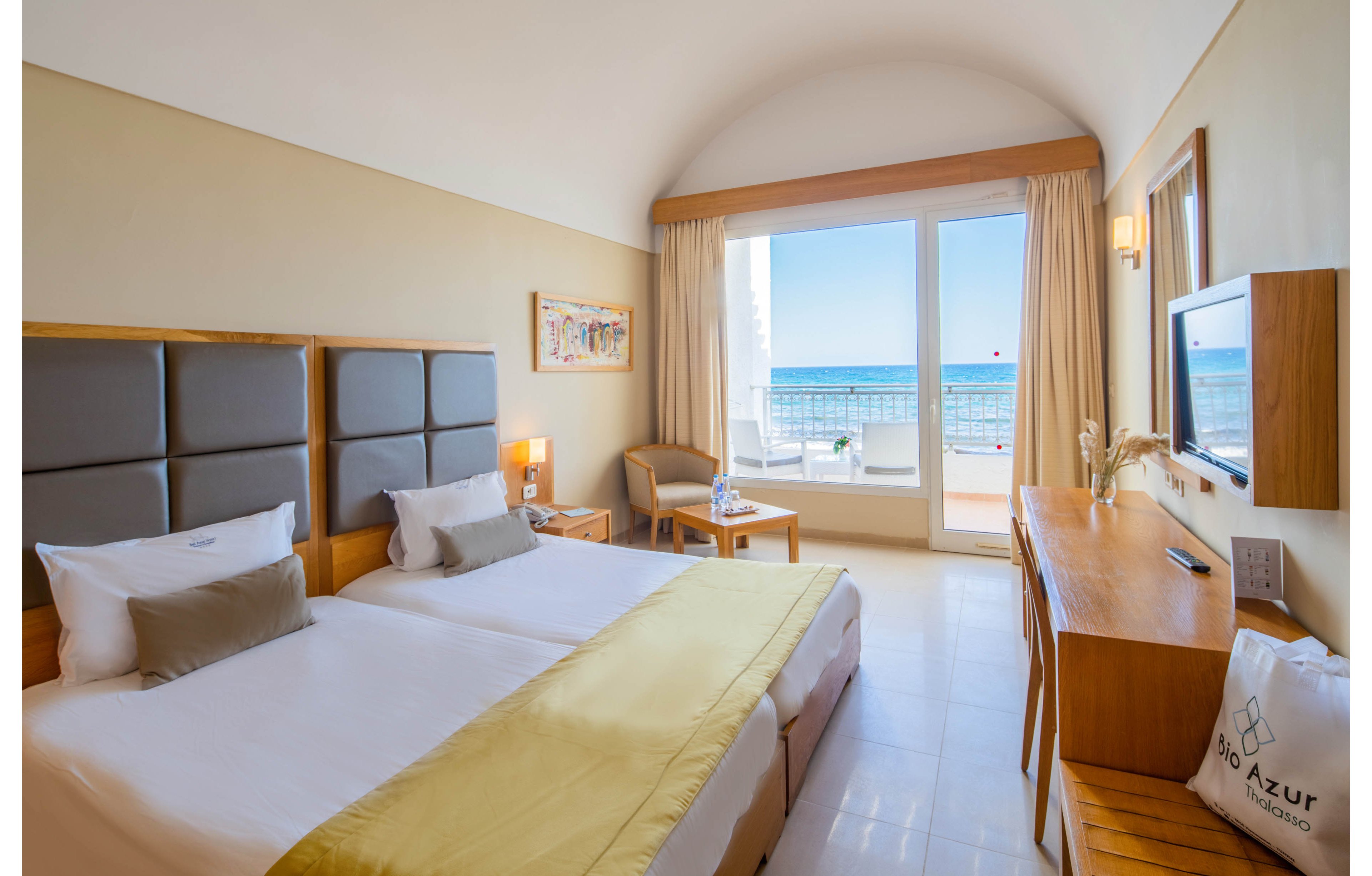 A modern hotel room features two single beds with neutral bedding and a soft yellow throw. The space includes a wooden desk, wall-mounted television, and a small seating area with a round table. Large glass doors open to a balcony overlooking a calm blue sea and clear sky, filling the room with natural light. Light-colored walls and minimal decor create a clean, airy atmosphere ideal for a beachside stay.