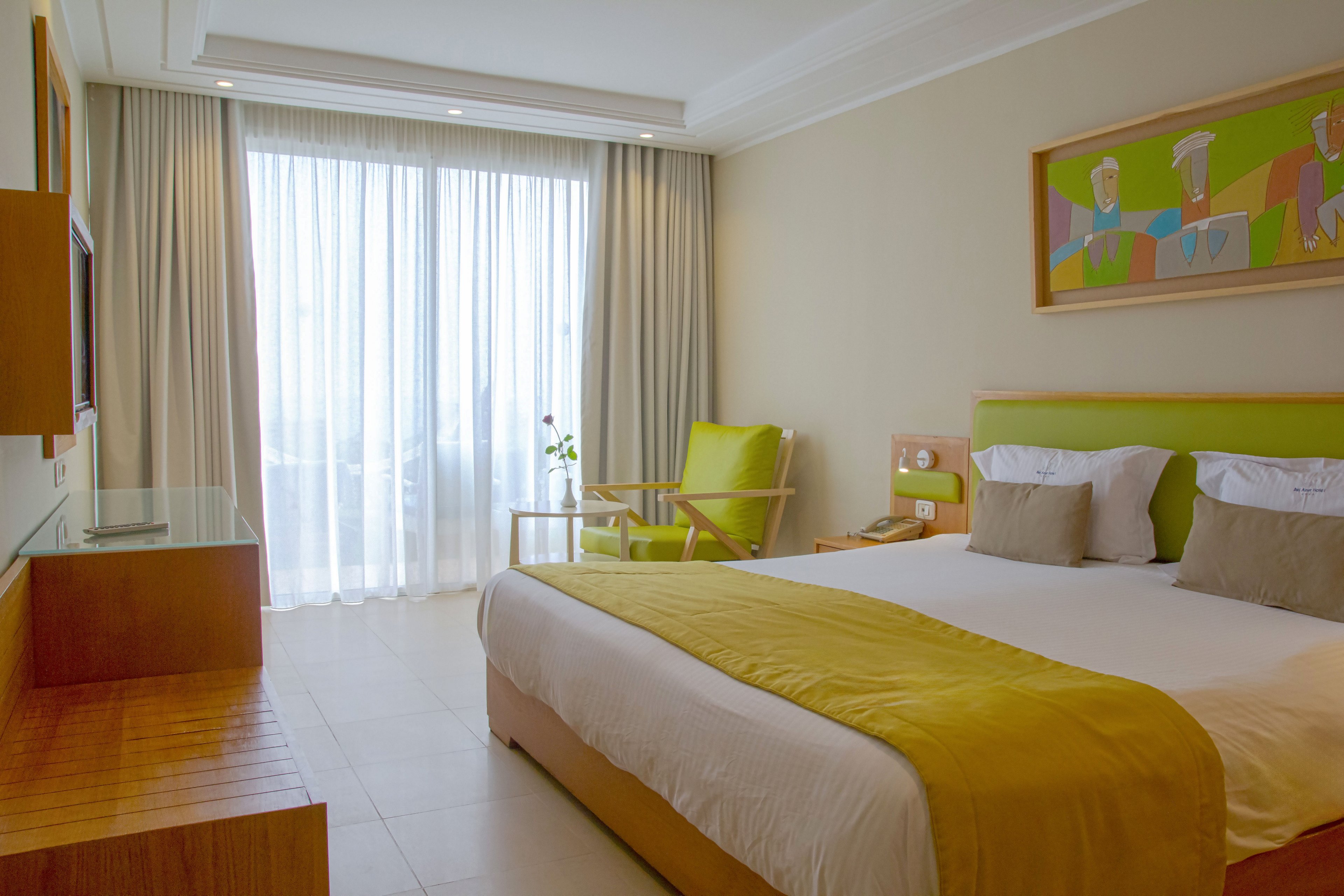 Contemporary hotel bedroom featuring a large double bed with white linens and green accents. The room is filled with natural light from a floor-to-ceiling window covered by sheer curtains and side drapes. A wooden desk, chair, and small side table create a simple seating and work area. Minimalist decor and a colorful abstract painting contribute to a clean, relaxing atmosphere.