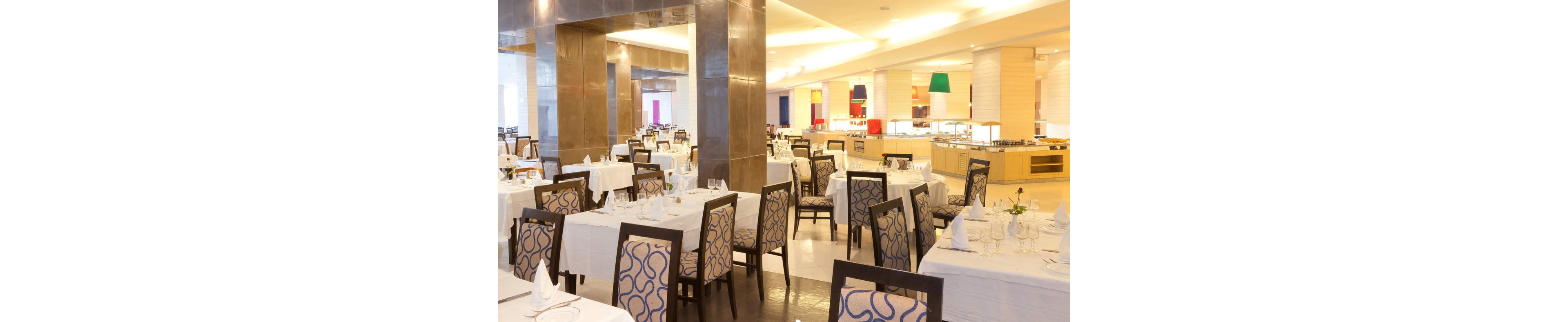Spacious contemporary restaurant dining area with neatly arranged tables and chairs. White tablecloths, folded napkins, and glassware are set for guests, but the room is currently empty. Warm ambient lighting and polished floors create an inviting atmosphere. Colorful accents and buffet counters are visible in the background, suggesting a hotel or resort setting.