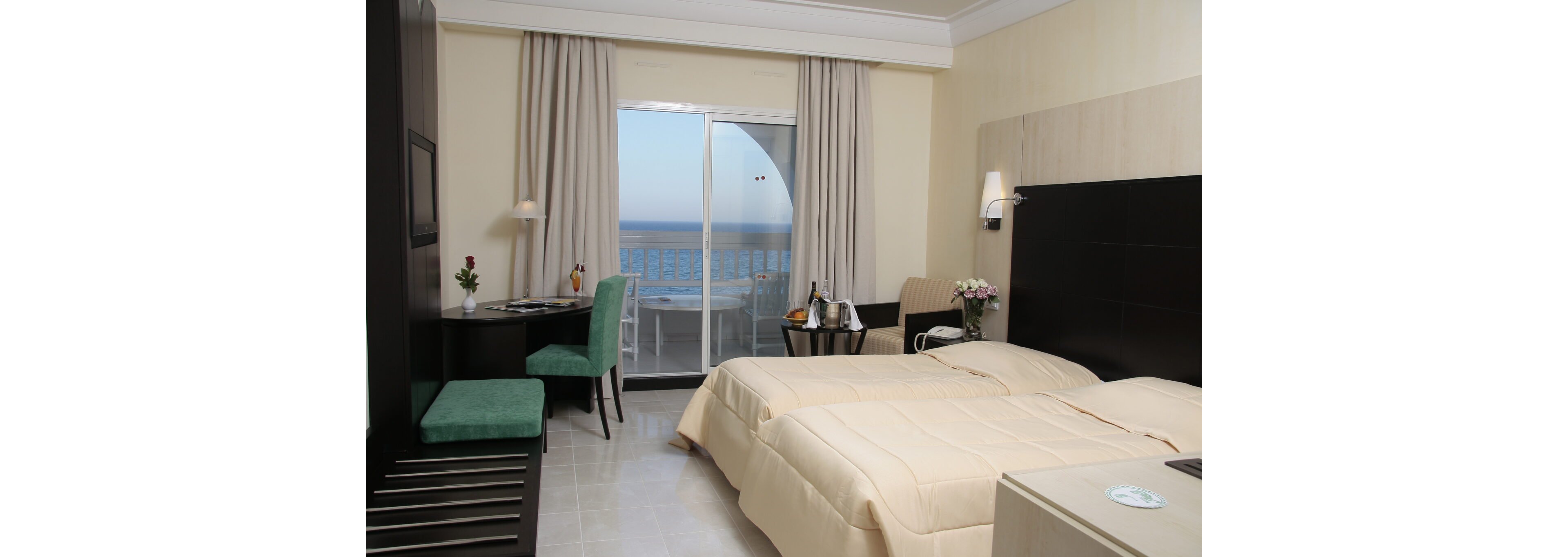 A contemporary hotel room features two neatly made twin beds with light beige bedding. A small desk with a green chair and wall-mounted television sits opposite the beds. Near the balcony doors, a table holds a wine bucket, glasses, and a vase of flowers, adding a touch of elegance. Large curtains frame the sliding glass doors that open to a balcony overlooking the ocean and clear sky.