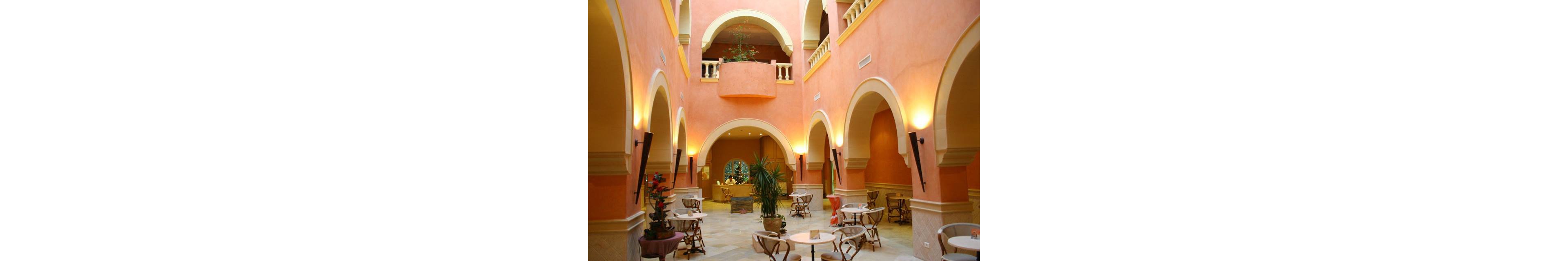 An elegant hotel atrium features peach-colored walls, high arches, and soft ambient lighting. Small round café tables with chairs are arranged across the marble floor, creating an inviting seating area. Potted plants and flowers add greenery and color to the open space. A reception or bar area is visible in the background beneath a large central archway.