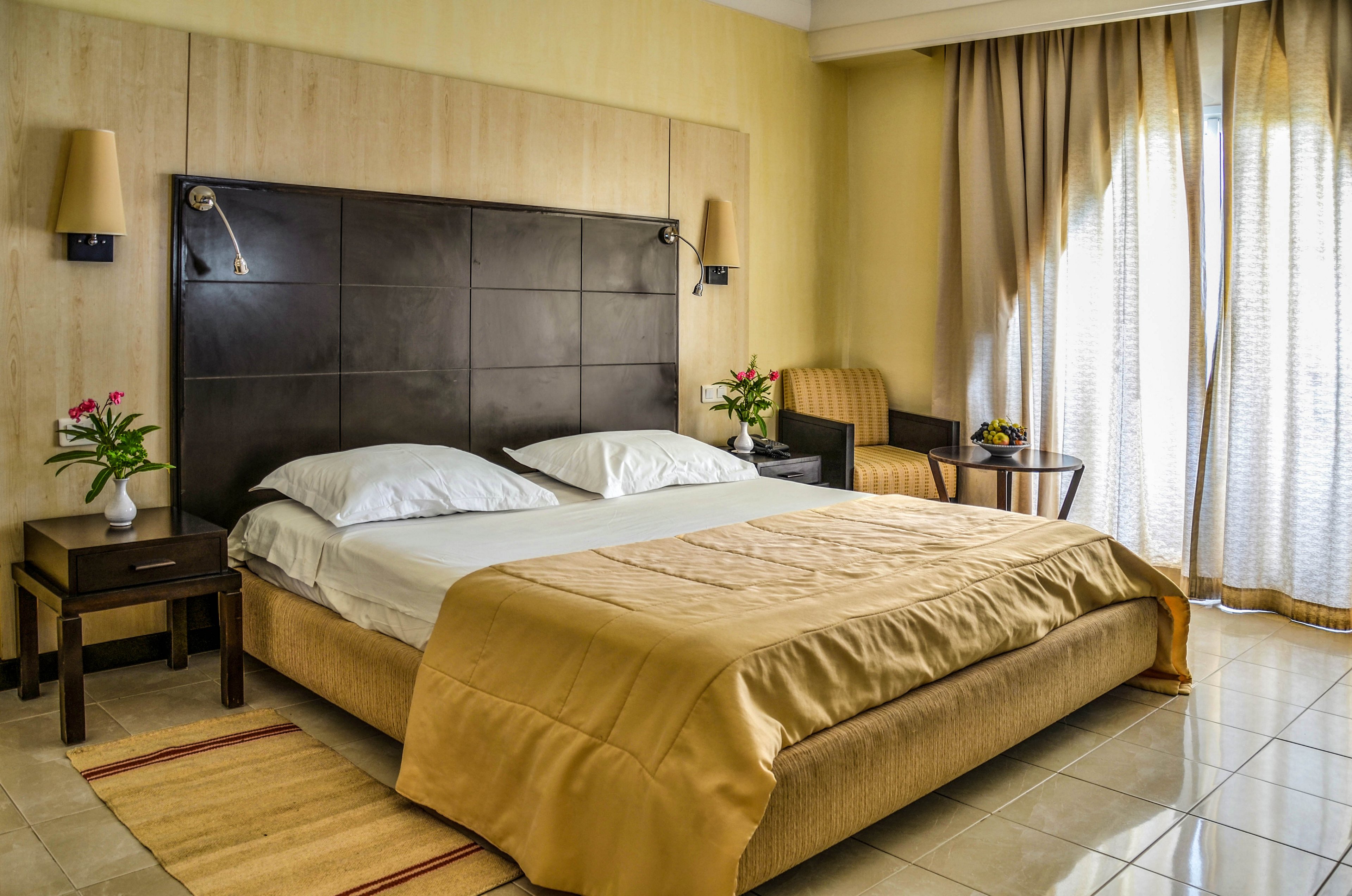 A neatly made double bed with white pillows and a beige bedspread is the focal point of this modern hotel bedroom. The room features matching bedside tables with lamps and small flower arrangements adding a touch of color. A striped armchair and small table sit near large windows covered with sheer and heavy curtains, allowing soft natural light to filter in. The overall design is clean and contemporary, with warm neutral tones and tiled flooring creating a calm, inviting atmosphere.