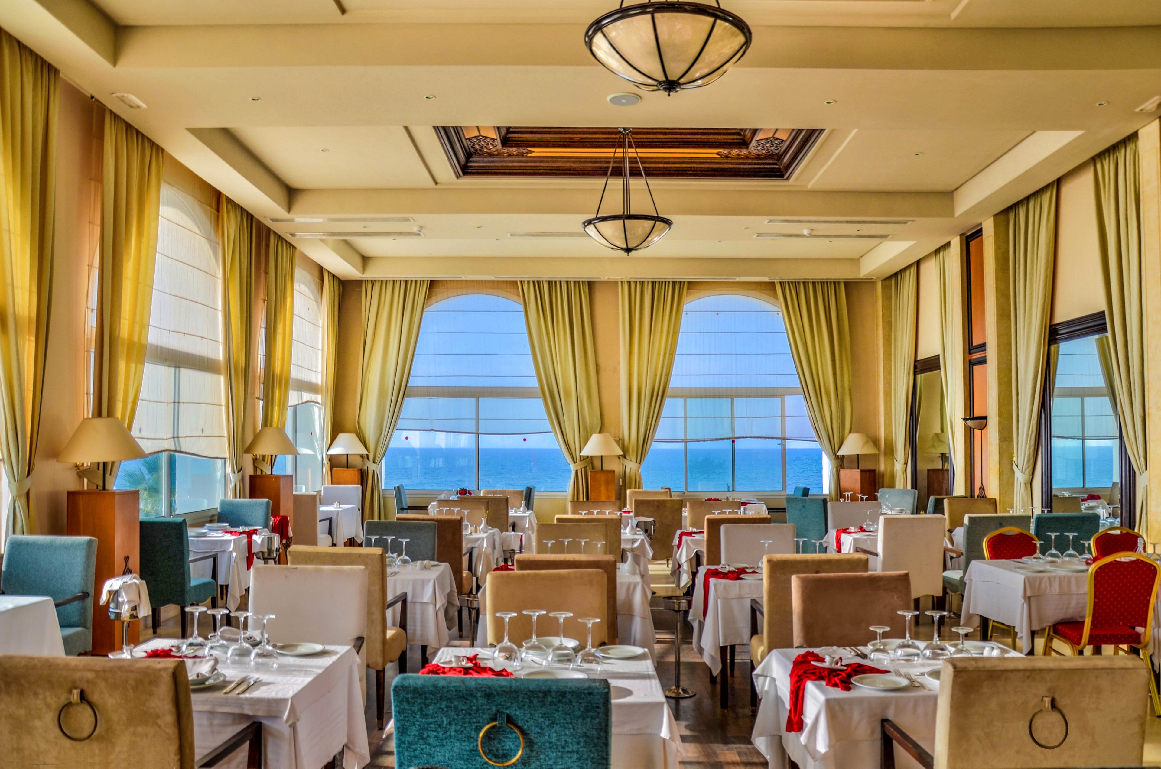 Upscale restaurant dining room with neatly arranged tables and chairs overlooks a bright seaside view through large windows. White tablecloths, folded napkins, and multiple wine glasses create a formal fine-dining atmosphere. Tall curtains frame the windows, while warm ceiling lights and chandeliers add a soft glow. The room is empty of people, emphasizing the symmetry, decor, and ocean scenery outside.
