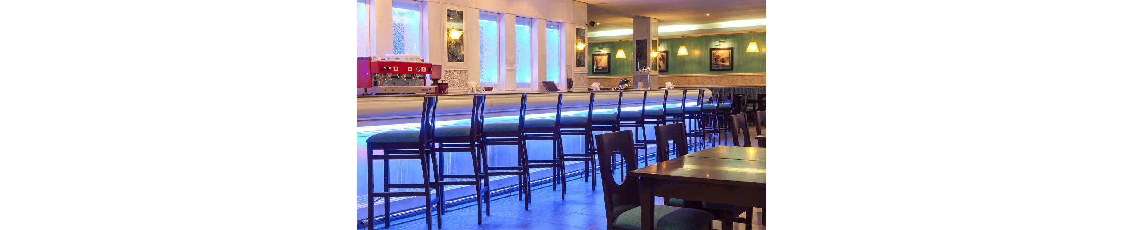 A long contemporary bar counter with high stools stands empty in a softly lit interior. Blue accent lighting runs along the bar front and windows, creating a cool, modern atmosphere. Pendant lamps and framed wall art add warmth to the otherwise sleek design. Dark wooden tables and chairs in the foreground suggest a restaurant or lounge setting ready for guests.