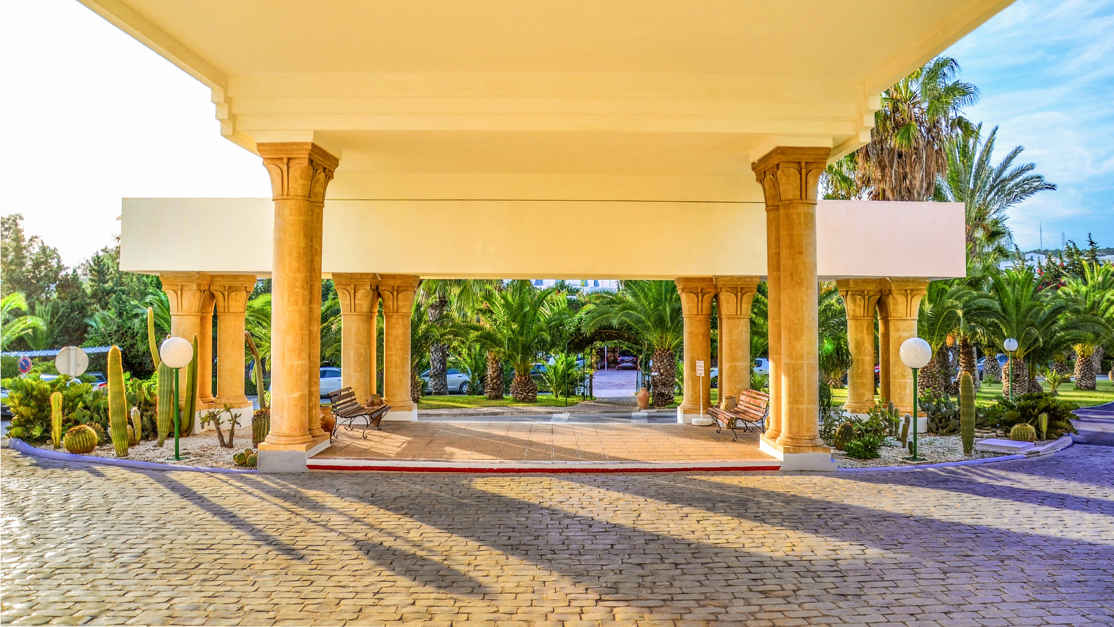 A wide, covered driveway leads to the entrance of a resort framed by tall columns. The foreground shows a cobblestone surface under the shade of the canopy. In the background, lush green palm trees and landscaped gardens are brightly lit by the sun. Benches and spherical pathway lights add to the welcoming, vacation-like atmosphere.
