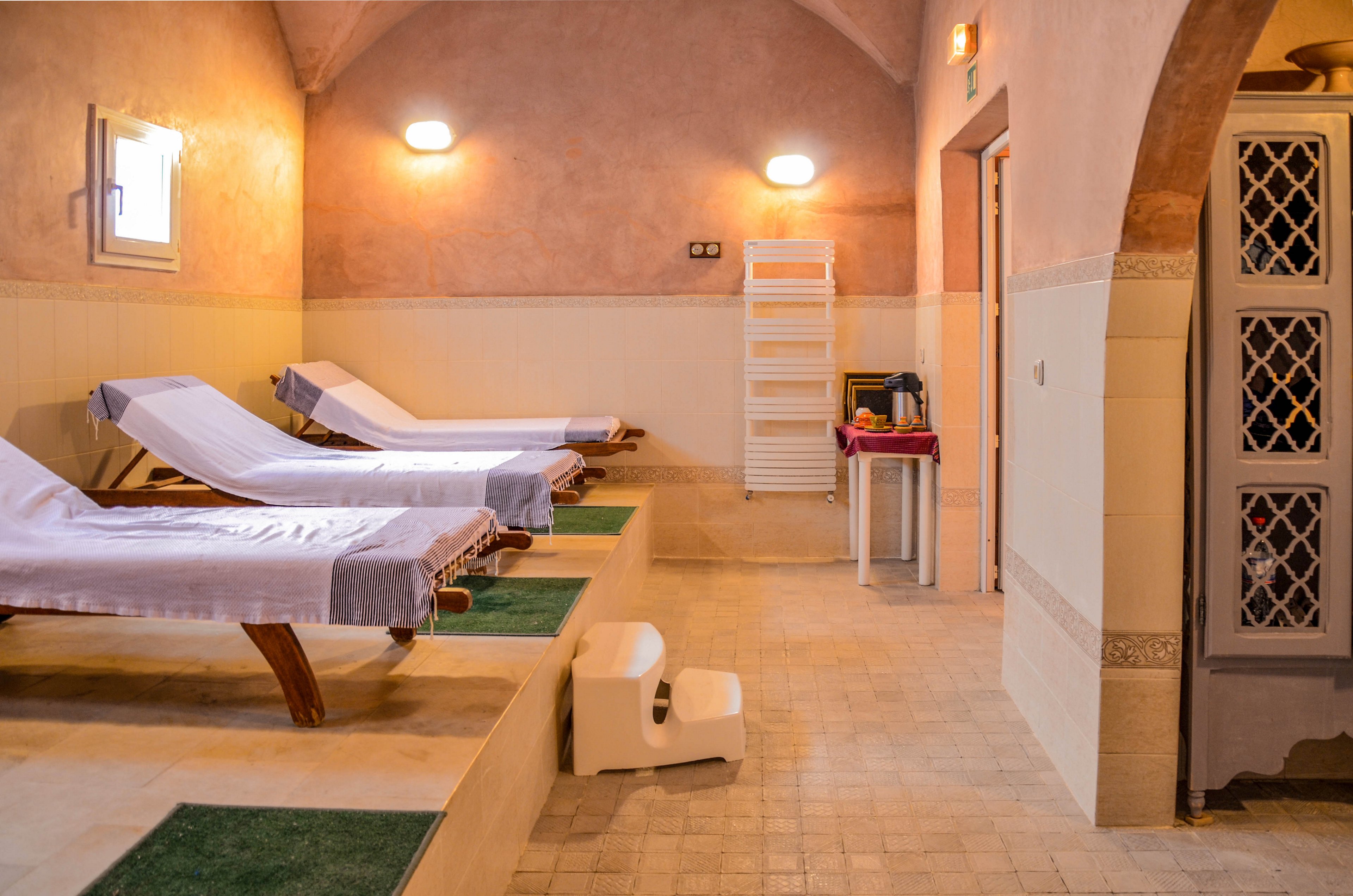 A serene spa relaxation room features several cushioned lounge chairs lined up on a raised tiled platform. Soft wall lighting and warm-toned plaster walls create a calm, inviting atmosphere. A small plastic step stool and a white towel rack stand nearby, adding functional details to the space. The overall setting suggests a peaceful wellness or spa environment designed for rest and recovery.