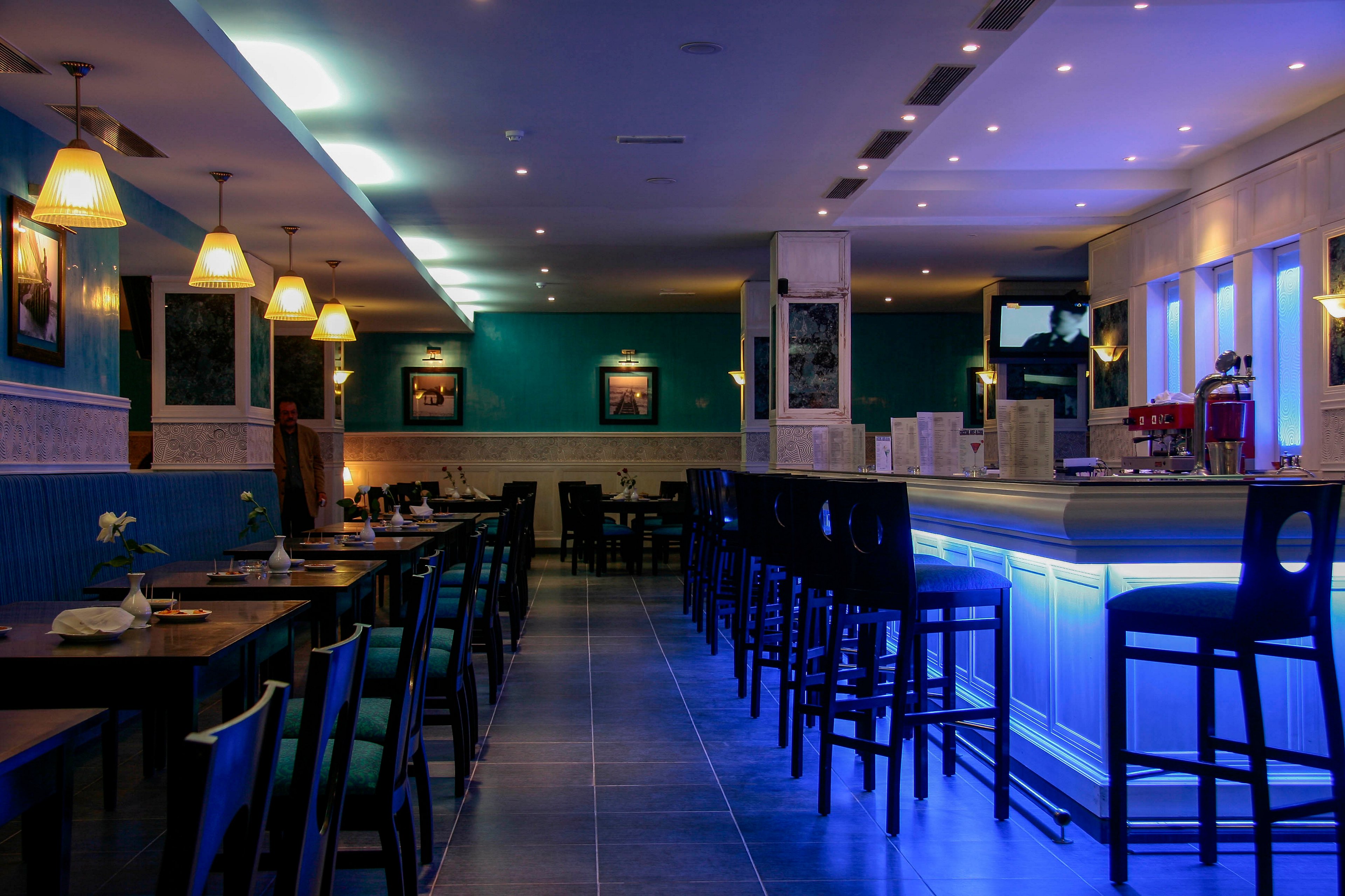 A contemporary restaurant interior features a long bar counter illuminated with cool blue lighting. Dark wooden tables and chairs line the left side, set neatly but currently unoccupied. Pendant lamps cast a warm glow over the seating area, contrasting with the blue accent walls and bar. Framed artwork and recessed ceiling lights add to the stylish, modern ambiance of the space.