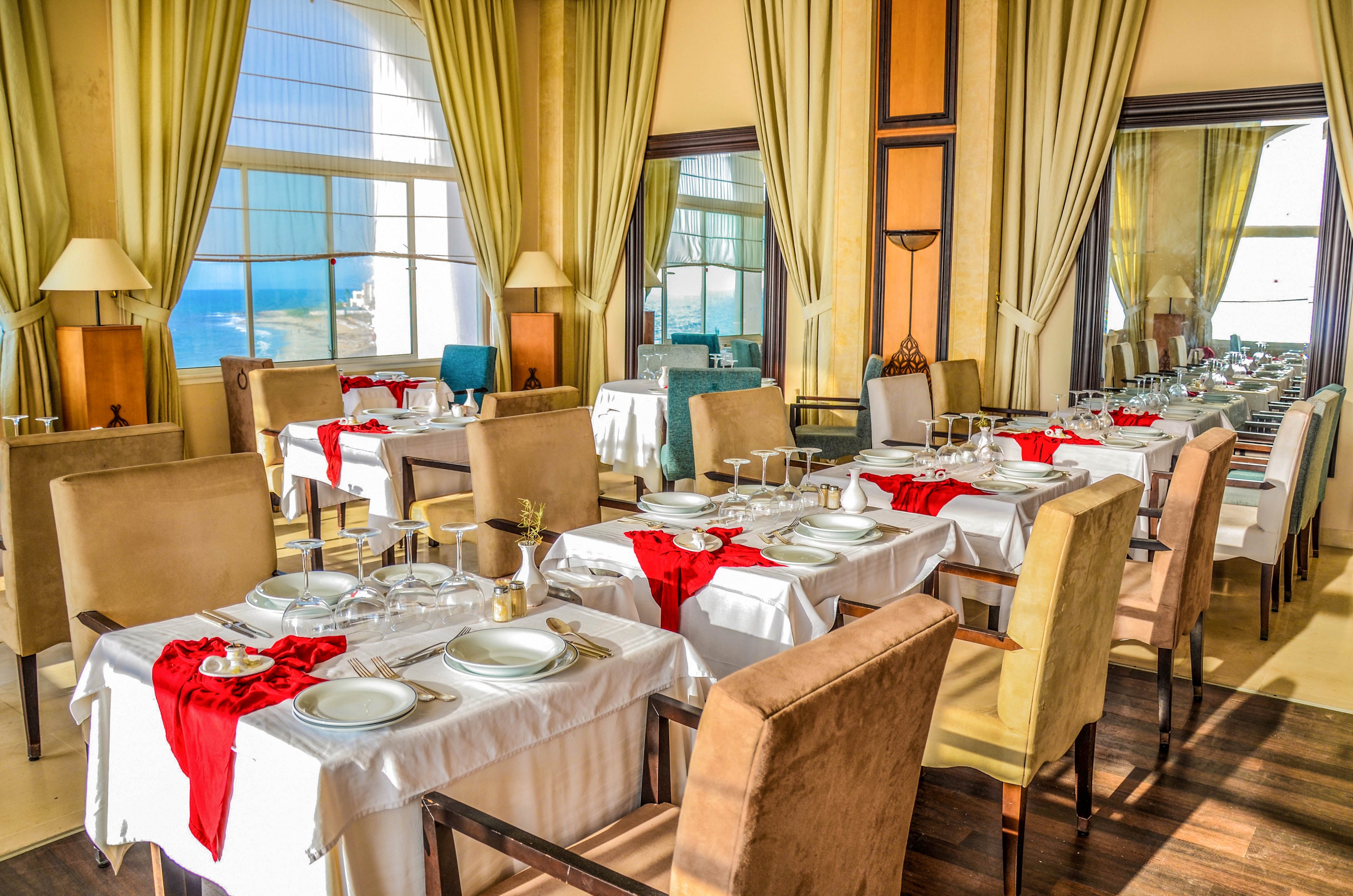A bright, upscale restaurant dining room is set with neatly arranged white tablecloths and formal place settings. Beige upholstered chairs surround the tables, which are accented with red napkins and glassware. Large windows and tall curtains allow abundant sunlight to flood the space and reveal a view of the sea outside. The overall atmosphere feels refined, inviting, and prepared for guests.