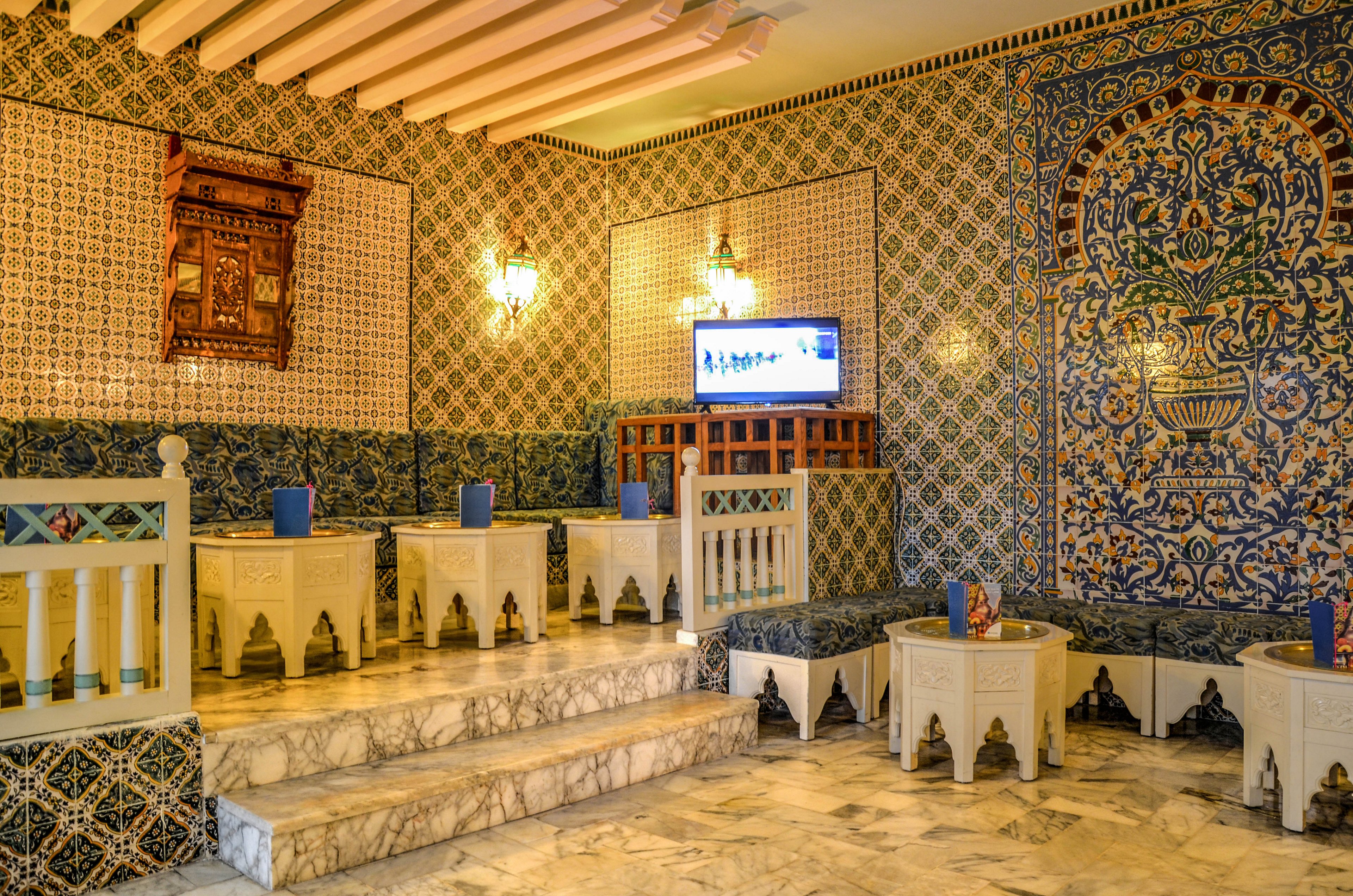 An ornate interior lounge features intricate patterned tiles covering the walls and benches. White carved wooden tables and railings sit on polished marble steps, creating a tiered seating area. Warm wall sconces illuminate the space, while a small television is mounted on the far wall. The overall atmosphere blends traditional craftsmanship with a touch of modern technology in a cozy, inviting setting.