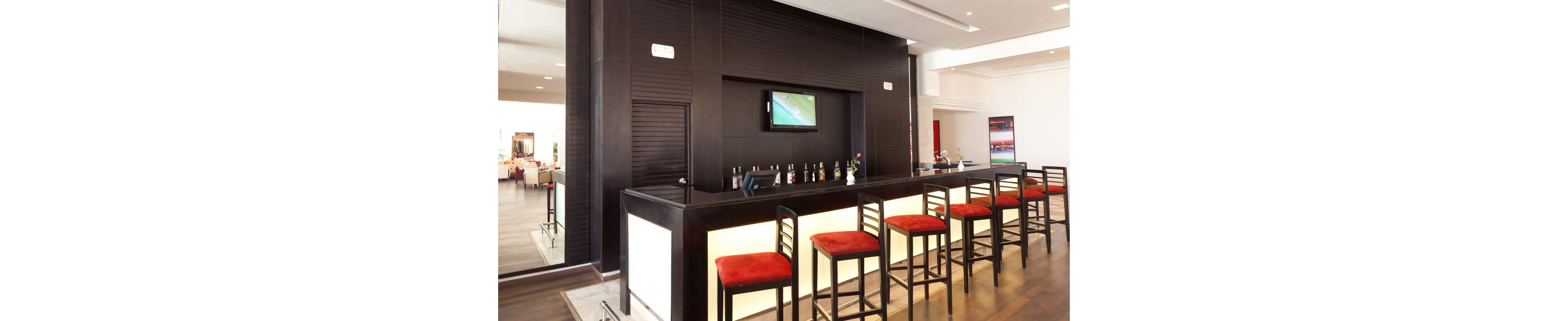 A sleek modern bar features a long counter with a glowing light panel and several red cushioned bar stools neatly aligned. Bottles of liquor and glasses are arranged behind the counter beneath a wall-mounted flat-screen television. The space is bright and minimal, with dark wood finishes contrasting against white walls and ceiling. In the background, additional seating and tables suggest a connected lounge or dining area.