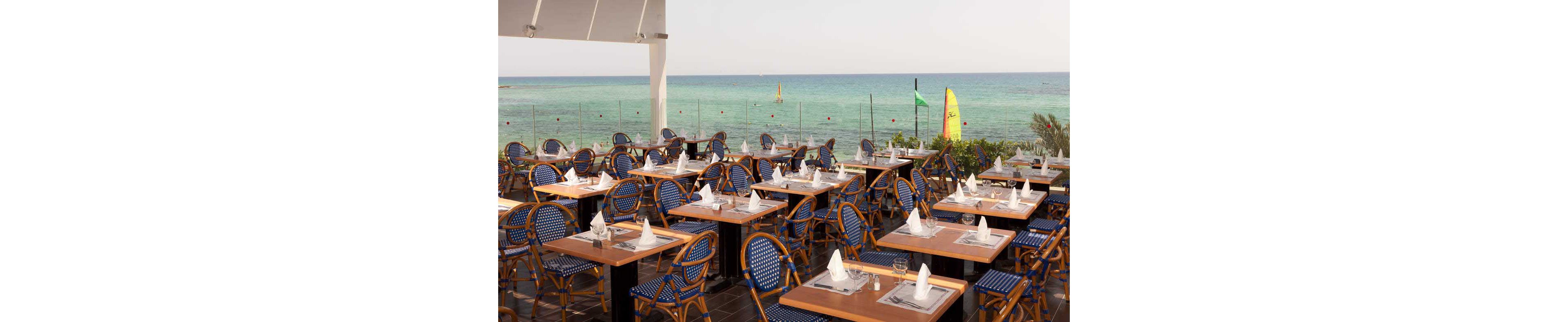 An outdoor restaurant terrace is set with neatly arranged wooden tables and blue woven chairs. Each table is prepared with white napkins folded into elegant shapes and glassware, ready for guests. Beyond the glass railing, the turquoise sea stretches to the horizon with small sailboats and a windsurfing board visible. The scene suggests a calm, sunny day at a coastal resort or beachfront dining area.