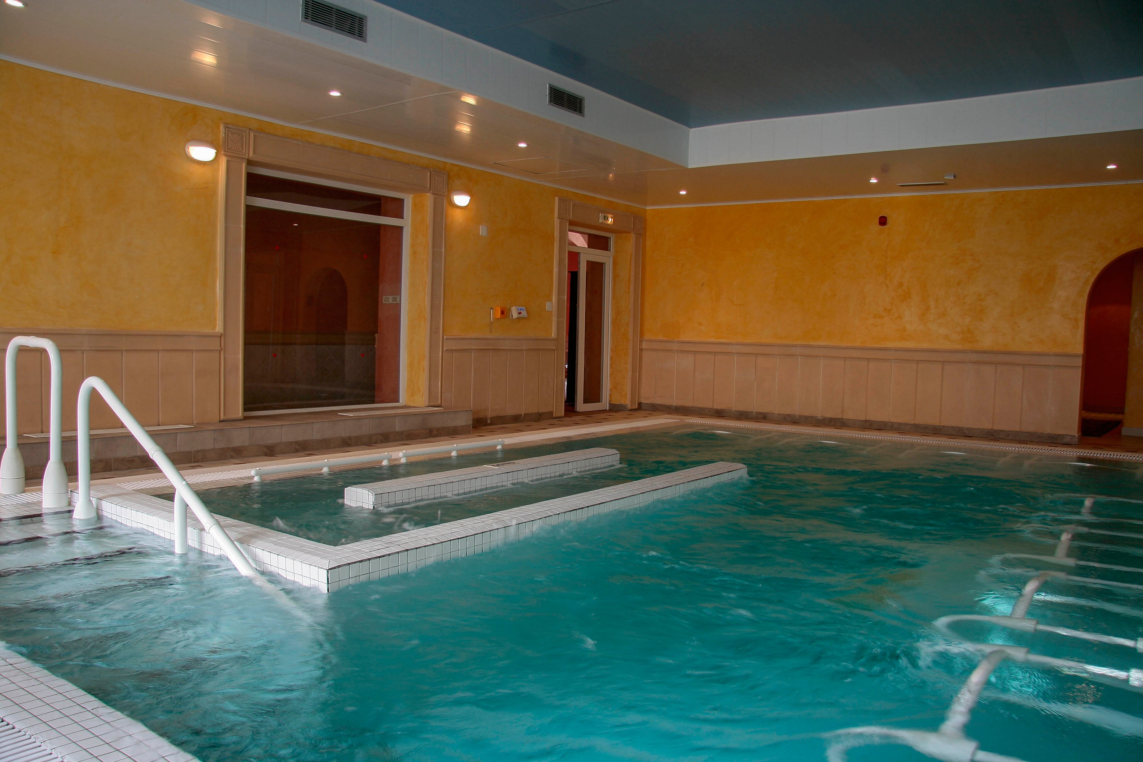 An indoor spa pool features clear blue water and multiple built-in sections for hydrotherapy. White railings and tiled steps lead into the pool, suggesting easy access for guests. Warm yellow walls and wood paneling create a relaxing, wellness-focused atmosphere. The space appears calm and empty, emphasizing tranquility and leisure.