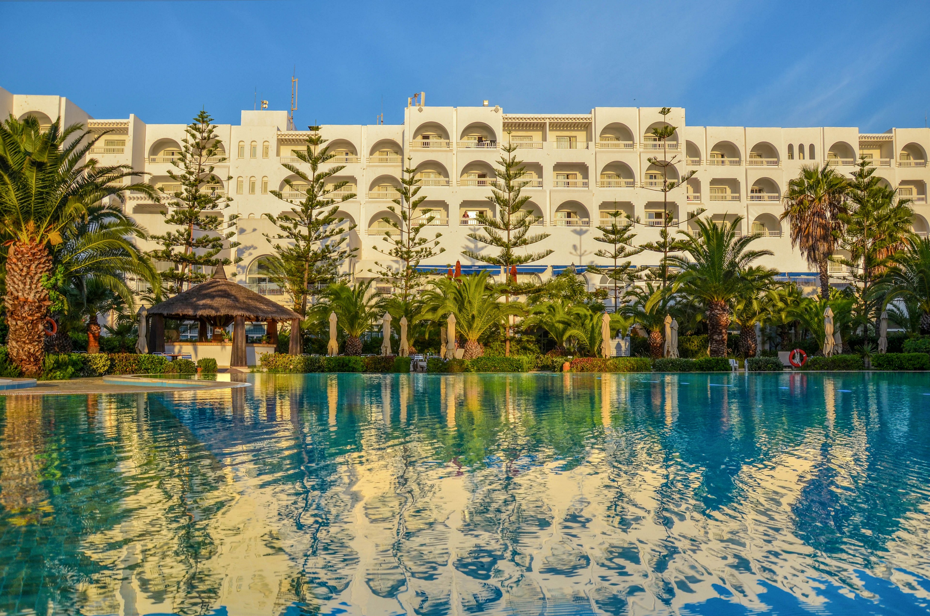 A large white resort hotel stands behind a tranquil outdoor swimming pool. Tall palm trees and manicured greenery line the edge of the water, creating a lush vacation setting. The calm pool surface reflects the building and trees in the warm golden light. Clear blue sky and soft evening or morning sunlight enhance the relaxed, tropical atmosphere.