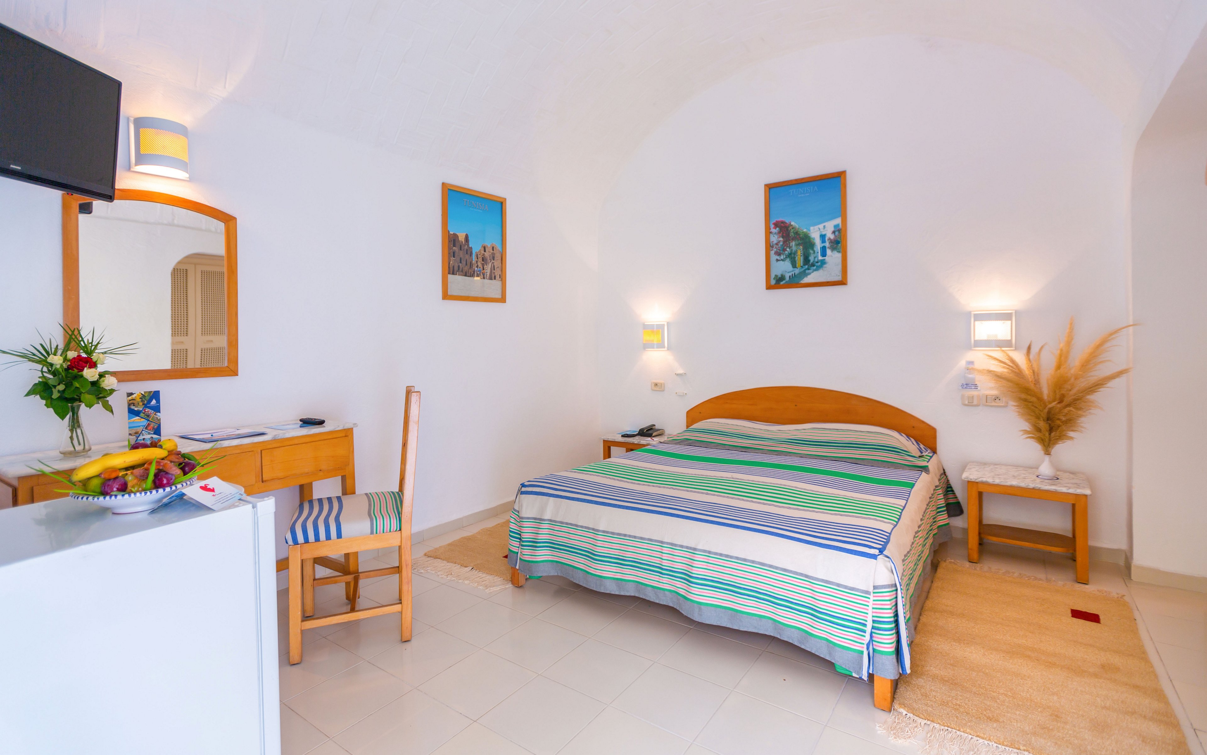 Cozy hotel bedroom with a double bed features striped bedding in soft blue and green tones. The room is decorated in a bright white Mediterranean style with arched ceiling, light wood furniture, and framed wall art. A desk with chair, fresh flowers, and fruit bowl add welcoming details, while bedside tables and wall lamps provide warm ambient lighting. Overall, the space feels clean, inviting, and ideal for a relaxing vacation stay.