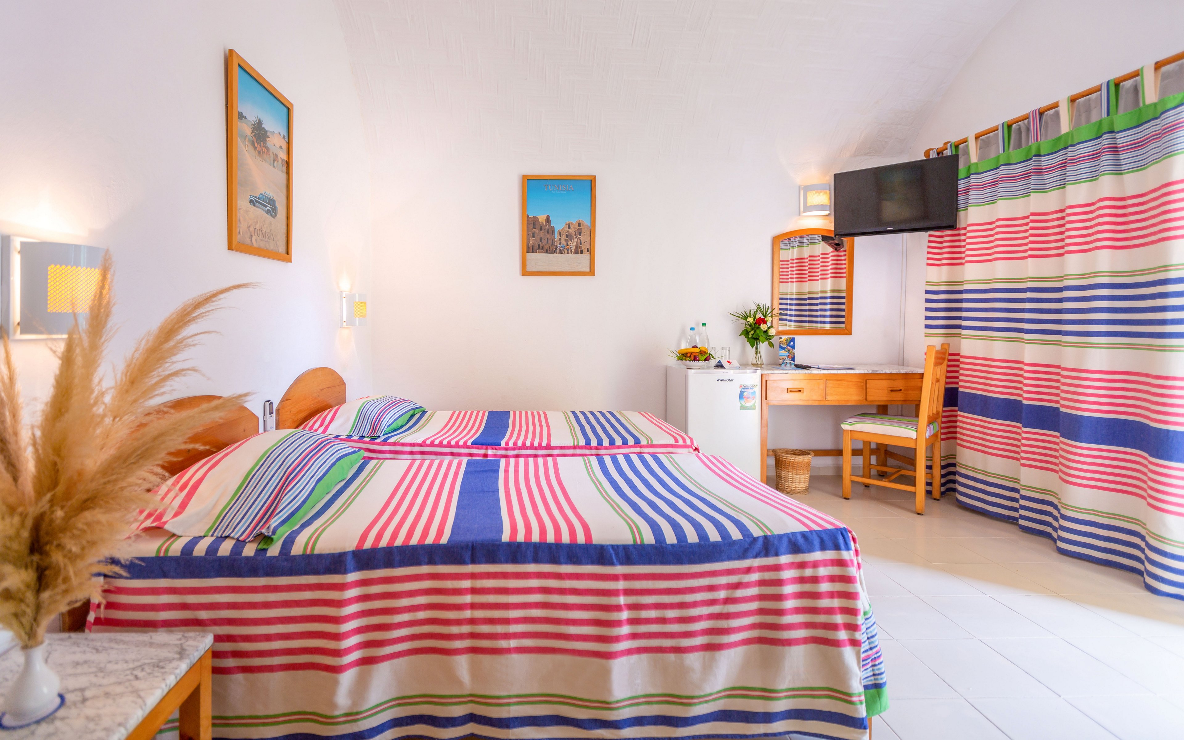 A colorful double bed with striped bedding is the focal point of a bright, airy bedroom. The room features white walls, framed artwork, and a wooden desk with a mirror and chair. A flat-screen television is mounted on the wall near vibrant striped curtains that match the bedspread. Decorative plants and dried grasses add a cozy, coastal touch to the clean, minimalist space.