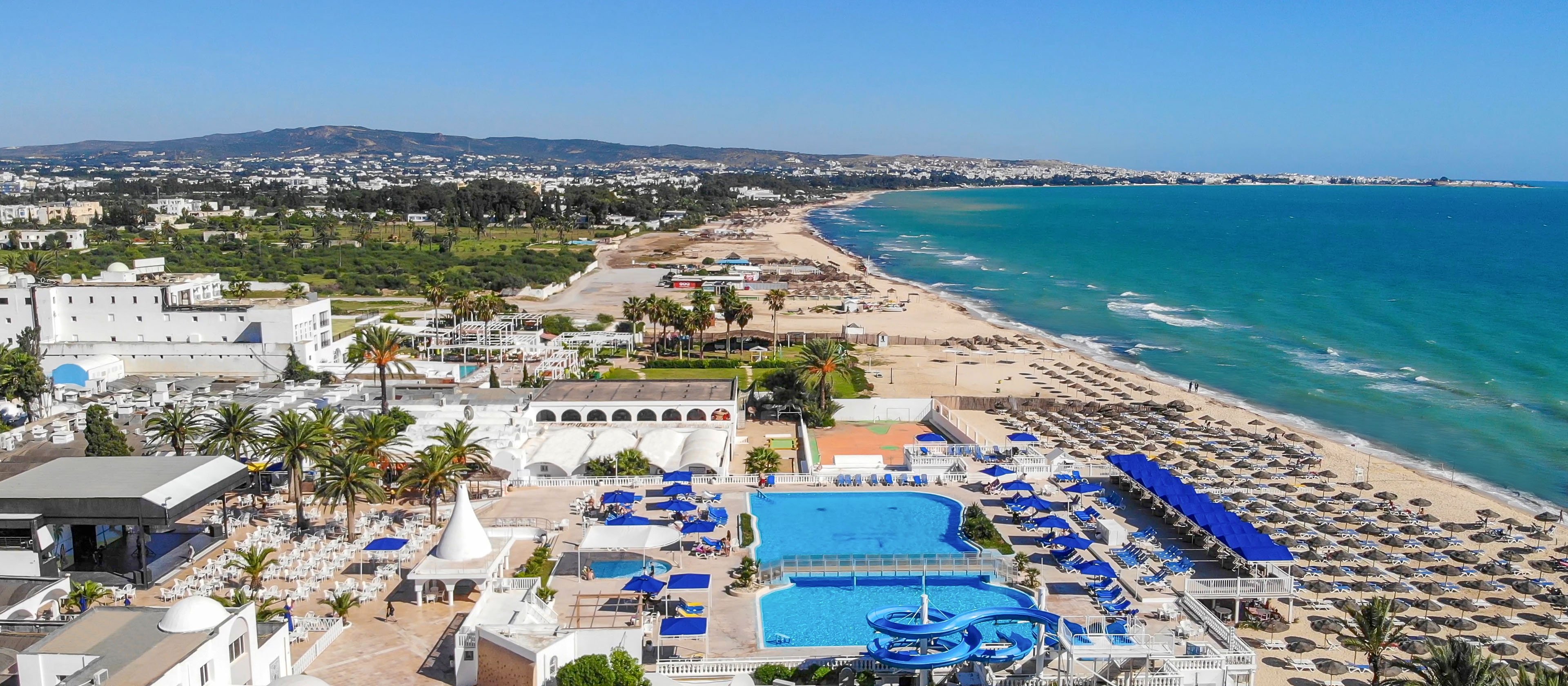 A large seaside resort stretches along a sandy coastline beside turquoise water. The complex features a central blue swimming pool surrounded by sun loungers and palm trees. Long rows of beach umbrellas line the shore, with more buildings and greenery extending into the distance. The scene is bright and sunny, suggesting a warm vacation destination on a clear day.