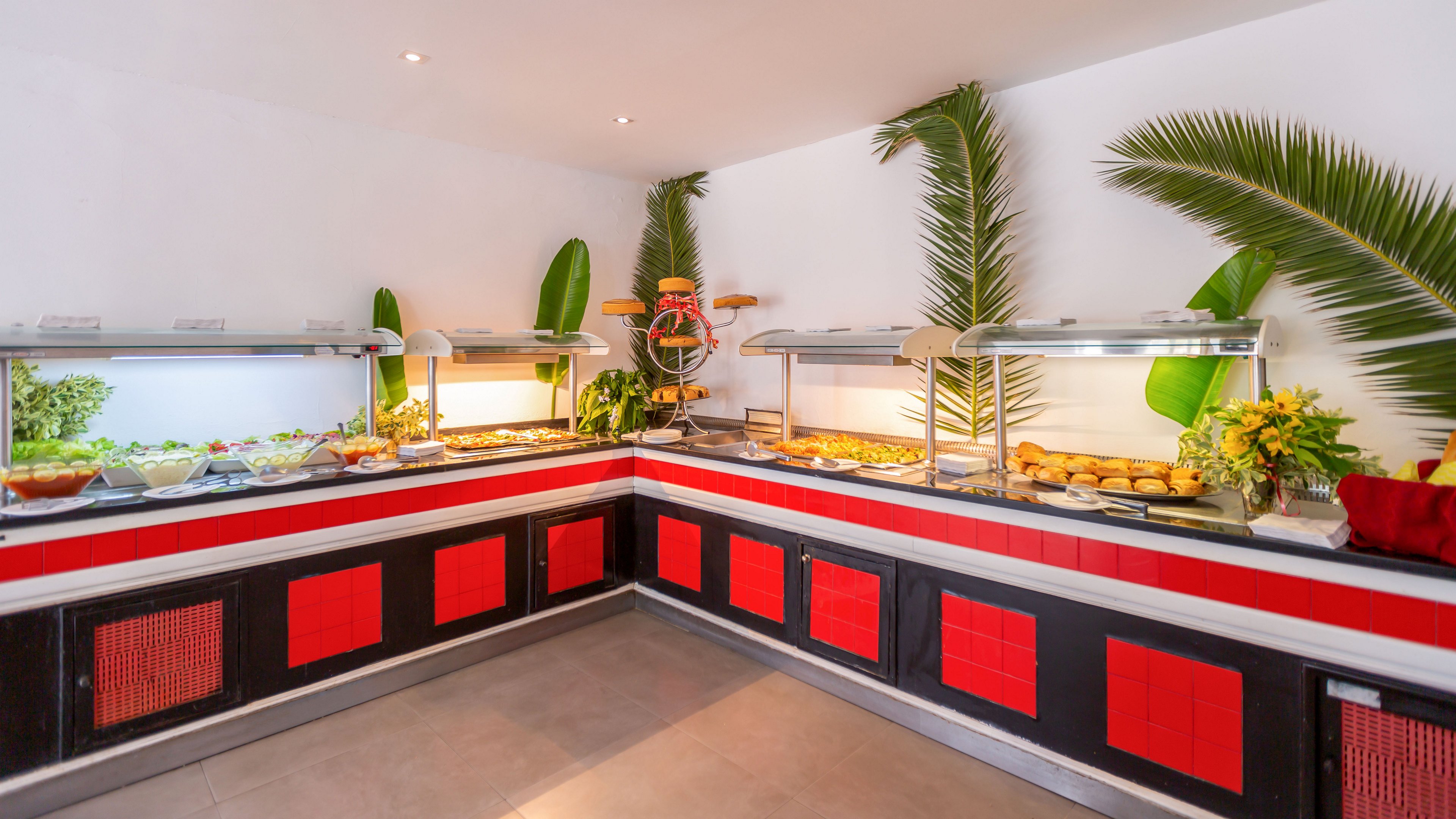 A corner buffet counter displays a wide variety of hot and cold dishes in stainless steel trays. The food is arranged under glass sneeze guards with warm lighting, suggesting a hotel or restaurant self-service area. Red and black panels decorate the counter base, while large green palm leaves and floral arrangements add a tropical touch. The clean tiled floor and white walls emphasize a neat, inviting dining environment.