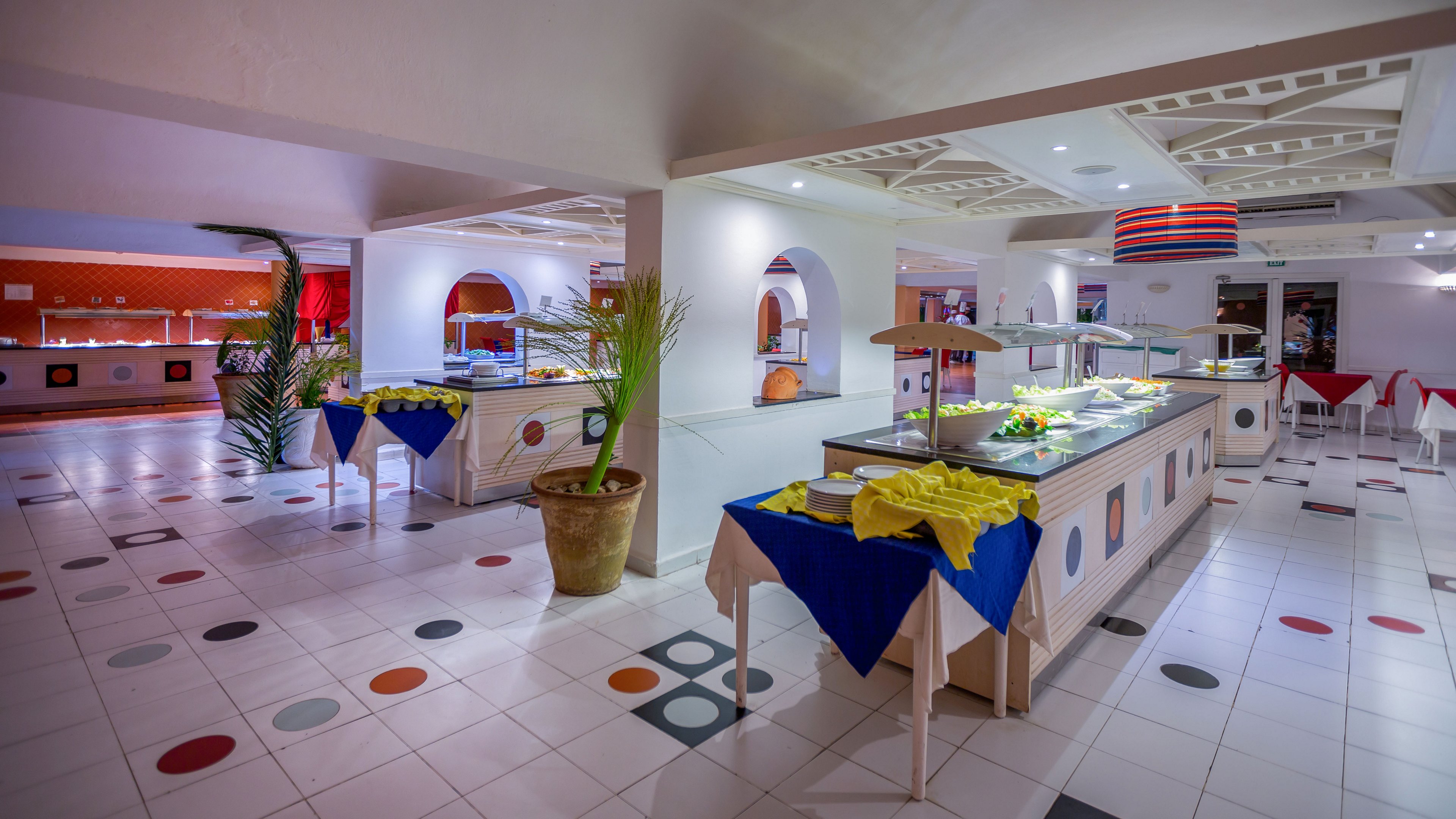 Spacious self-service buffet counters stand in a brightly lit hotel dining area. Tables are covered with blue and yellow cloths, and decorative plants in pots add greenery to the tiled floor. The white ceiling and floor feature geometric patterns with circular accents in multiple colors, creating a playful visual style. Food trays and serving stations are arranged neatly, suggesting a prepared but currently quiet restaurant setting.