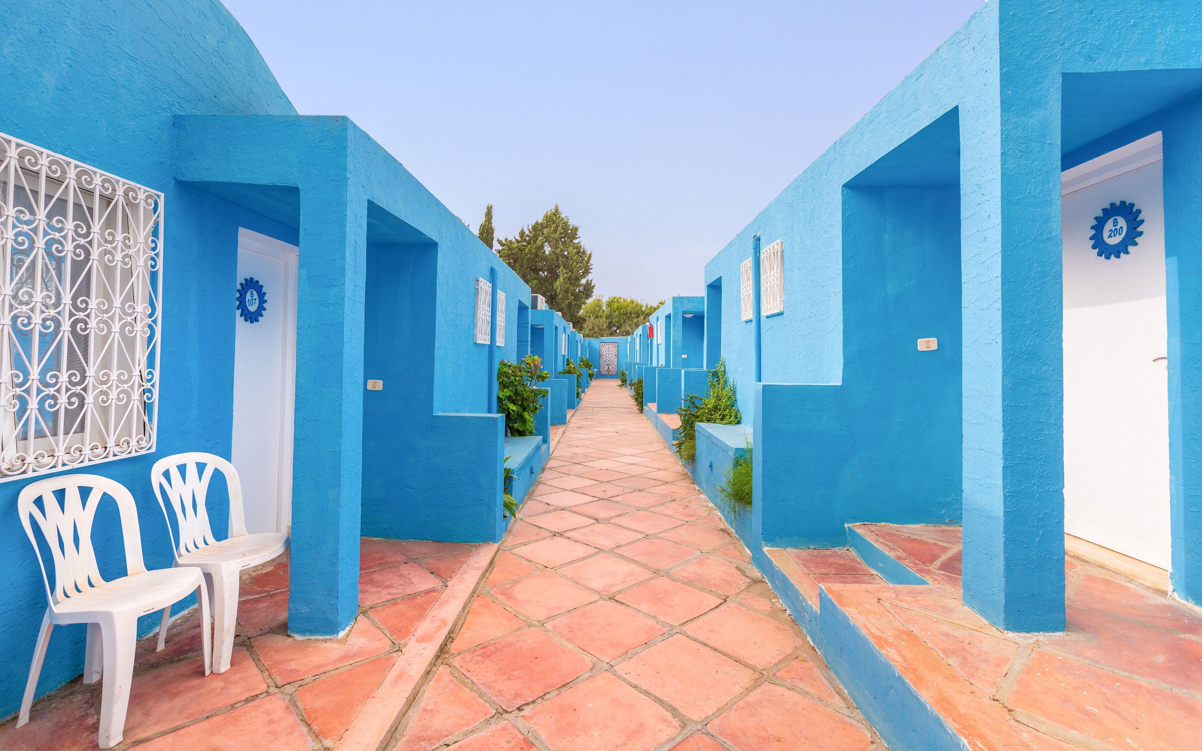 A narrow courtyard is lined with vivid blue buildings on both sides, creating a strong geometric perspective toward the distance. White doors and decorative metal grilles contrast with the blue walls, while potted plants add touches of green along the walkway. Two white plastic chairs sit near one doorway, enhancing the casual, relaxed atmosphere. The terracotta-colored tiled path leads the eye through the scene under a clear, pale sky.
