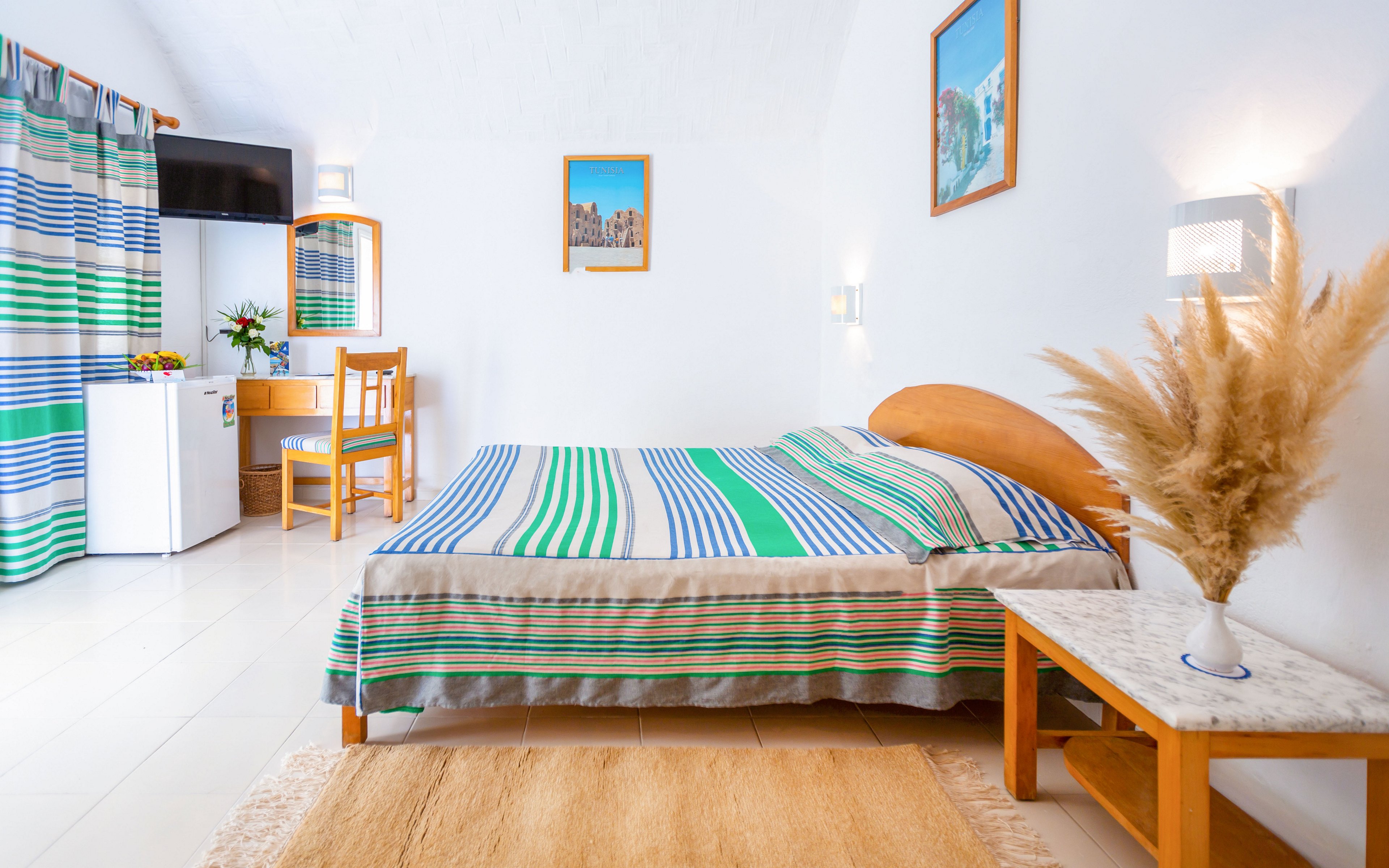 A tidy bedroom features a double bed with a colorful striped bedspread in blue, green, and white tones. The room is decorated with matching striped curtains, light wooden furniture, and framed wall art. A small desk area with a chair, television, and mini fridge sits in the corner, while a side table holds a vase of dried pampas grass. The white walls and ceiling, along with the neutral rug, create a bright and airy coastal atmosphere.