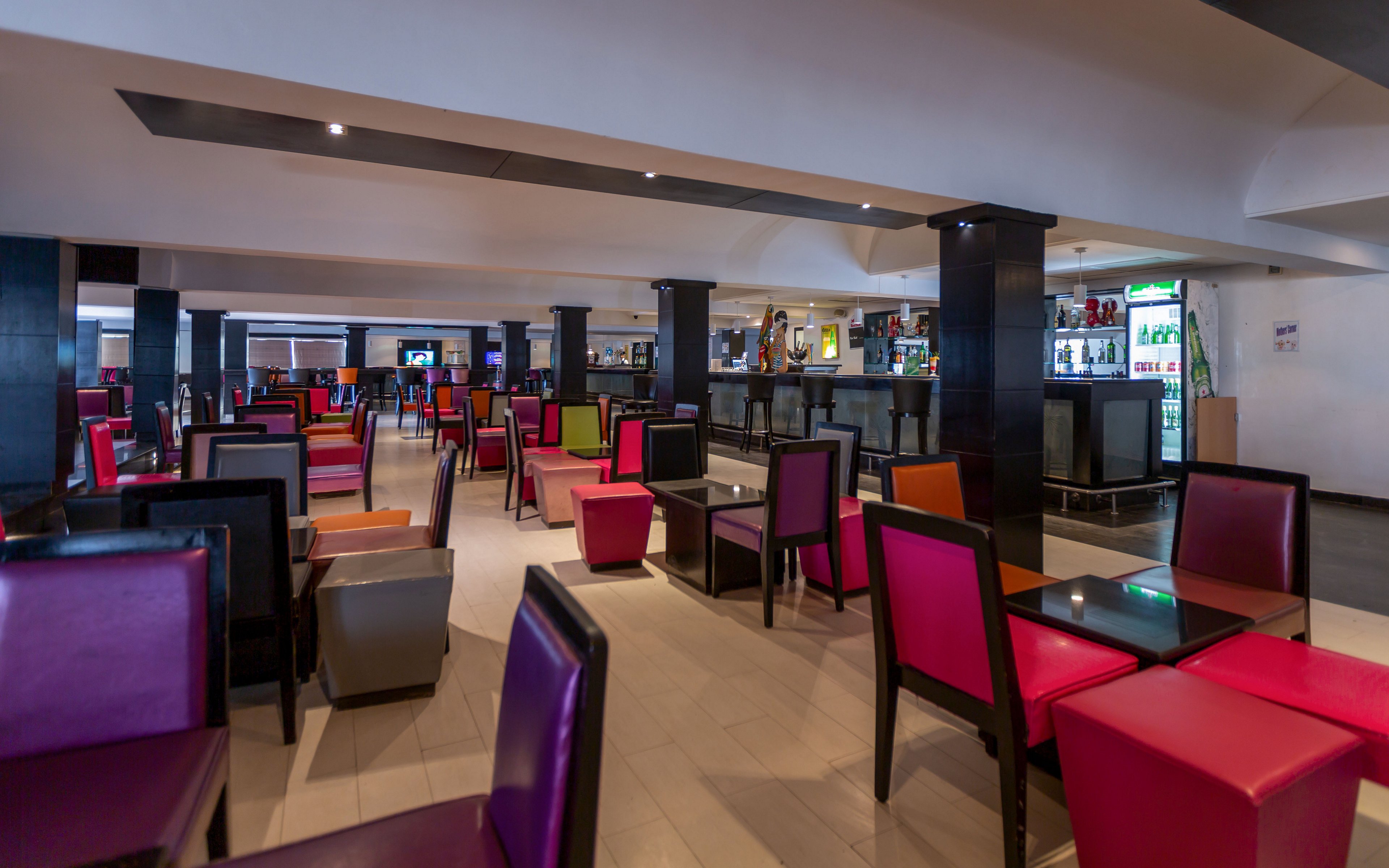The image shows a spacious, modern restaurant interior with rows of tables and chairs. Brightly colored upholstered chairs in red, pink, purple, and green create a vibrant atmosphere against neutral floors and walls. A long bar counter with bottles and glassware is visible in the background, along with refrigerated display units. The space appears clean, organized, and ready to receive guests.