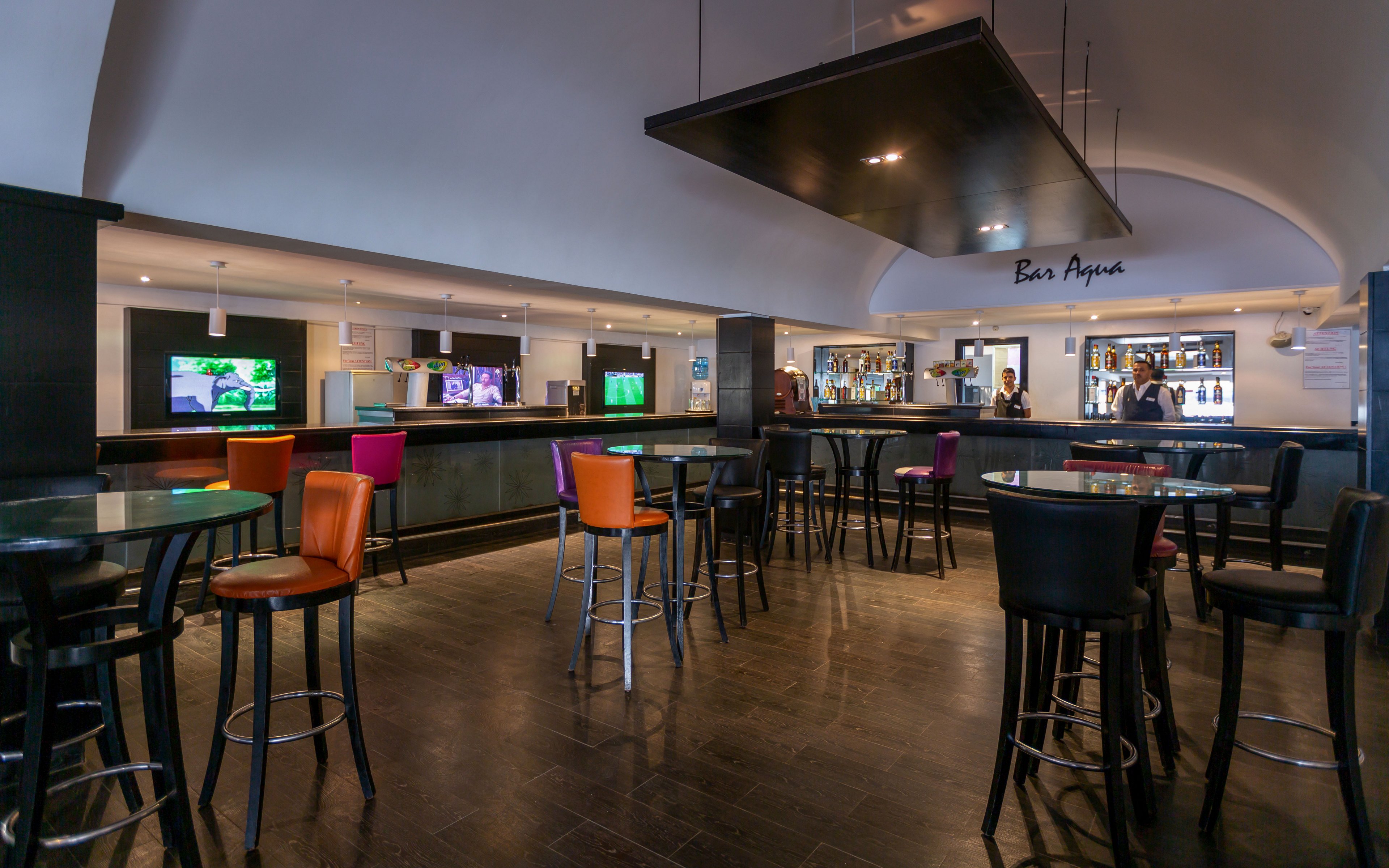 Spacious contemporary bar interior with high tables and colorful stools arranged across a dark tiled floor. The long counter at the back features illuminated shelves, drink displays, and multiple television screens showing various programs. A few people stand near the bar area, adding a casual social atmosphere to the otherwise open space. The arched ceiling and sleek lighting fixtures create a modern, inviting setting suitable for nightlife or hotel lounge scenes.