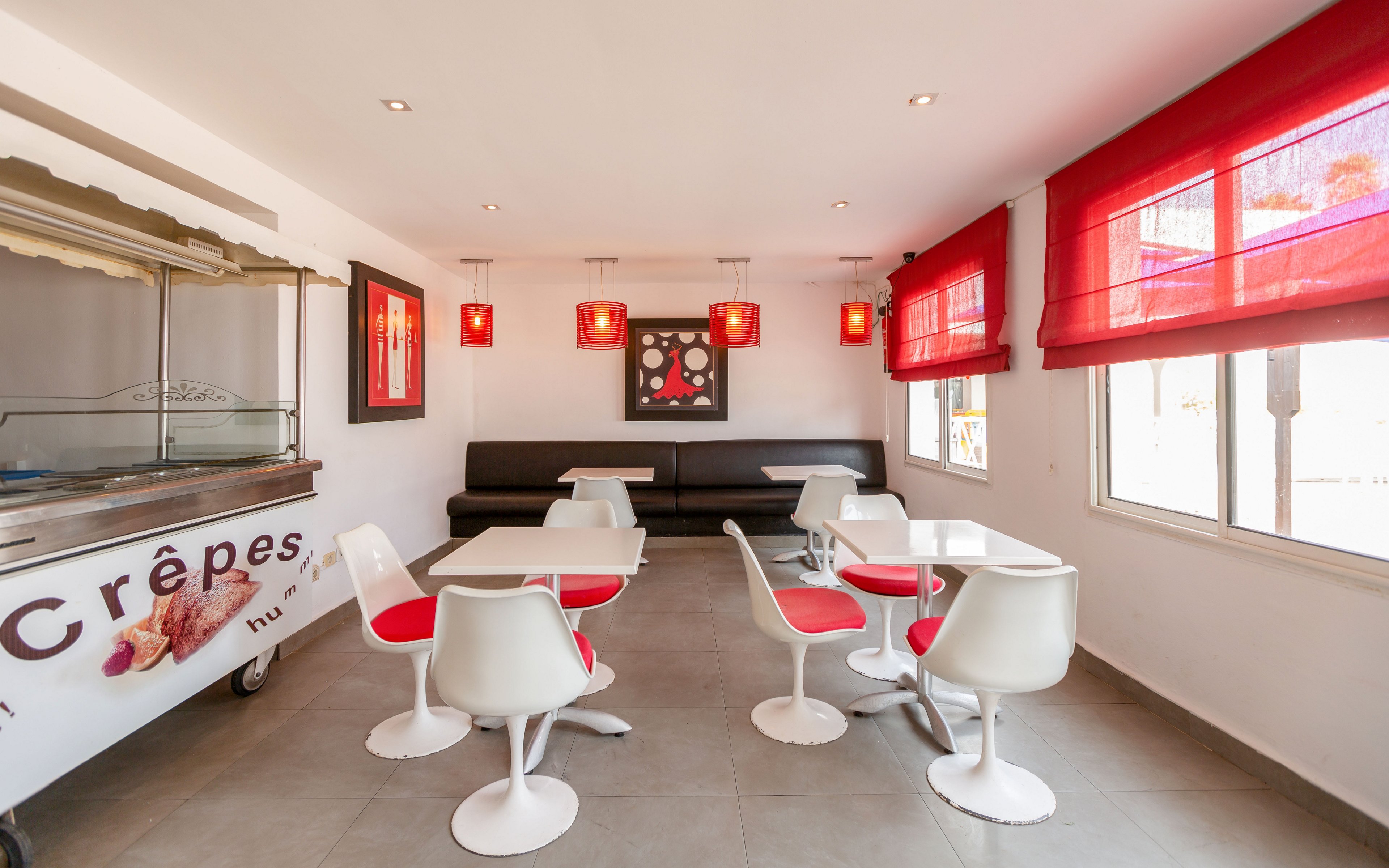 A small modern café interior features white tables with red and white chairs arranged neatly in the center of the room. On the left side, a crepe stand with the word “Crêpes” and an image of crepes is visible. Red pendant lights and red window blinds contrast with the otherwise white walls and ceiling, creating a bold color scheme. Large windows allow natural light to fill the space, highlighting the clean, minimalist design and empty seating area.
