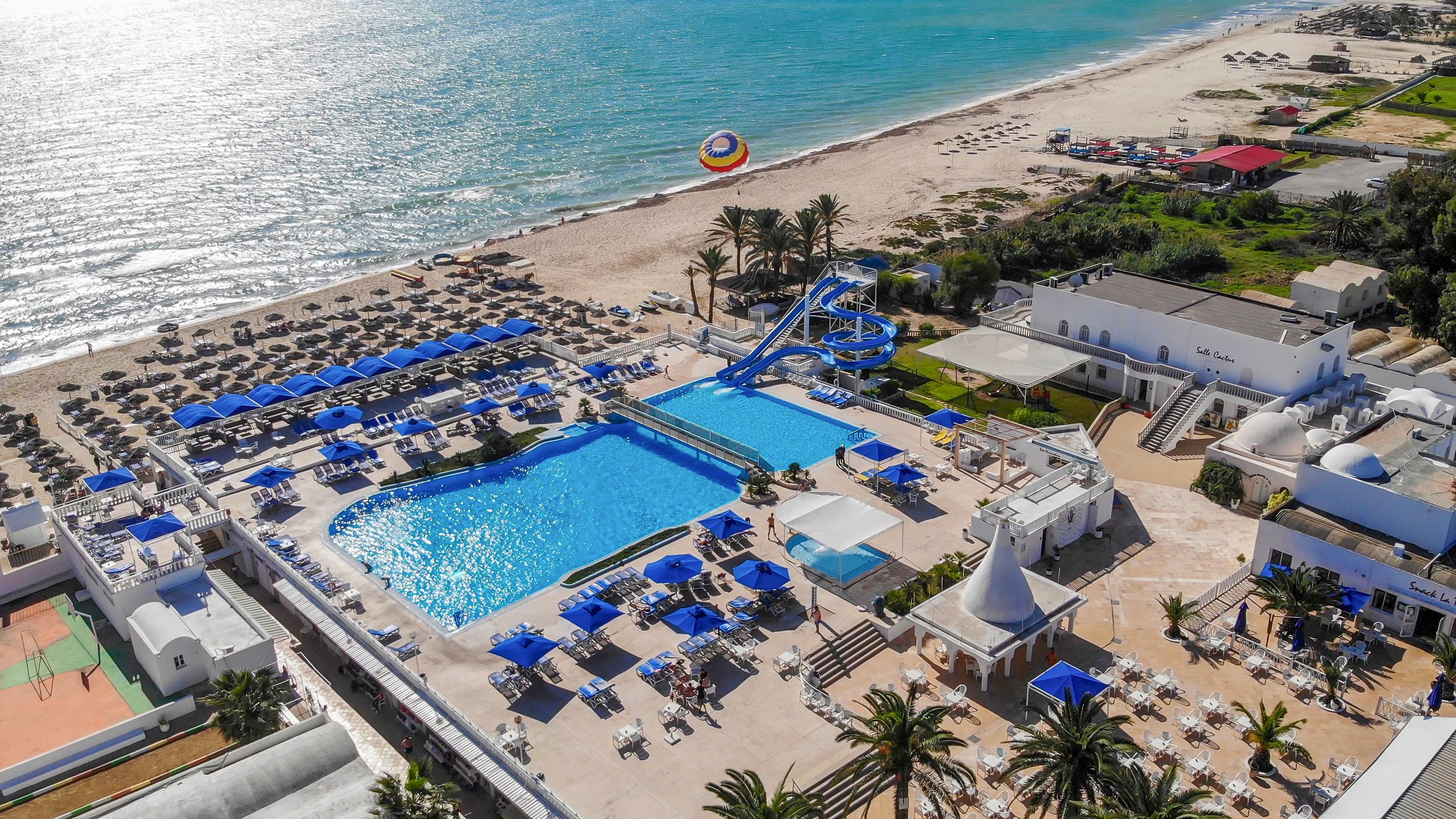 A large beachfront resort features a central blue swimming pool surrounded by sun loungers and umbrellas. Water slides lead into an adjacent pool area, adding a sense of fun and activity. The property sits directly on a sandy beach lined with rows of deck chairs facing the ocean. Bright blue accents contrast with white buildings and terraces, creating a sunny Mediterranean holiday atmosphere.