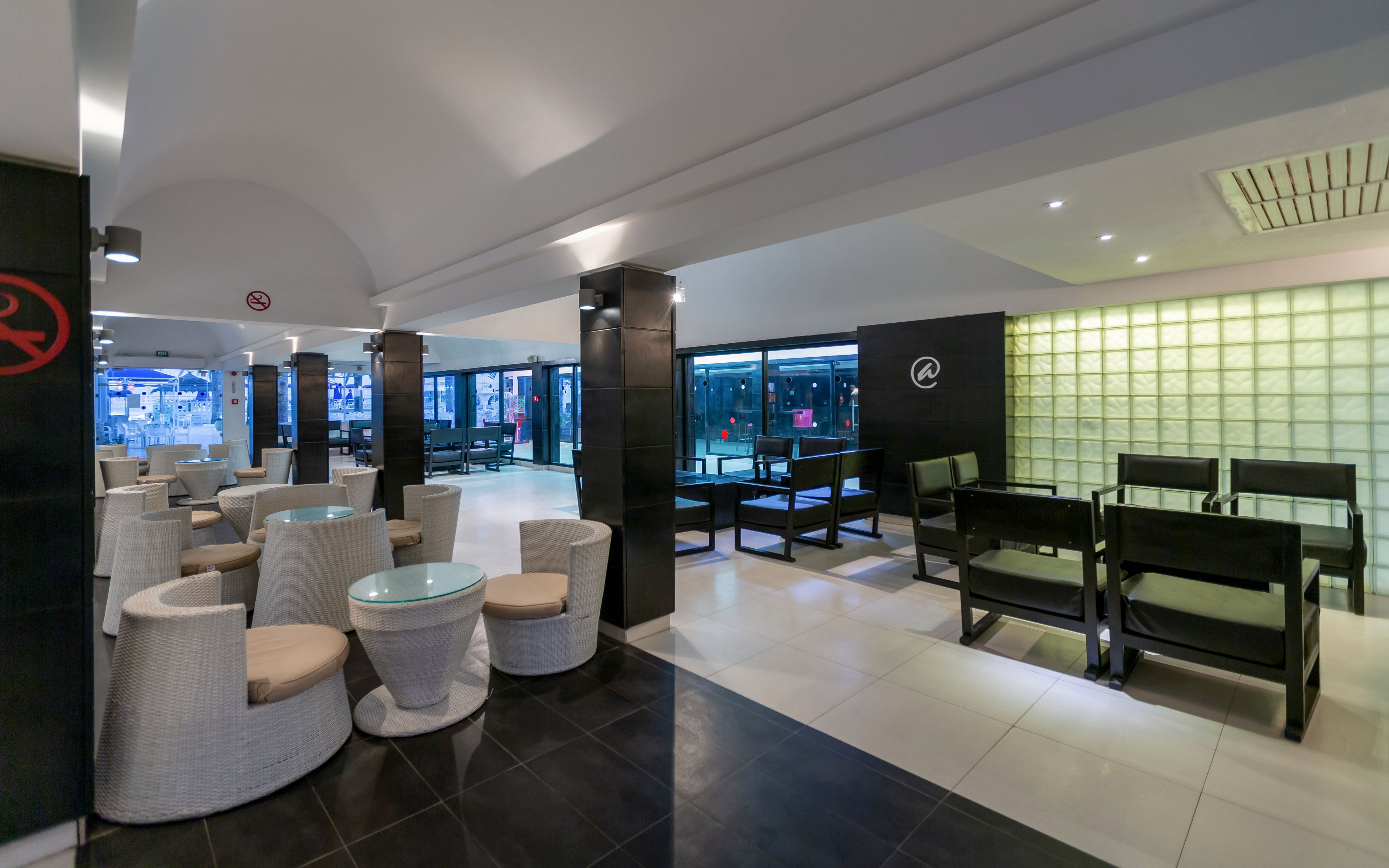 Contemporary lounge interior featuring both round wicker chairs with glass-top tables and dark wooden dining-style seating. The space is open and spacious, with tiled flooring and a white arched ceiling supported by dark columns. Large windows line one side, offering views of the exterior and natural light. A backlit glass block wall adds a stylish, modern accent to the room.