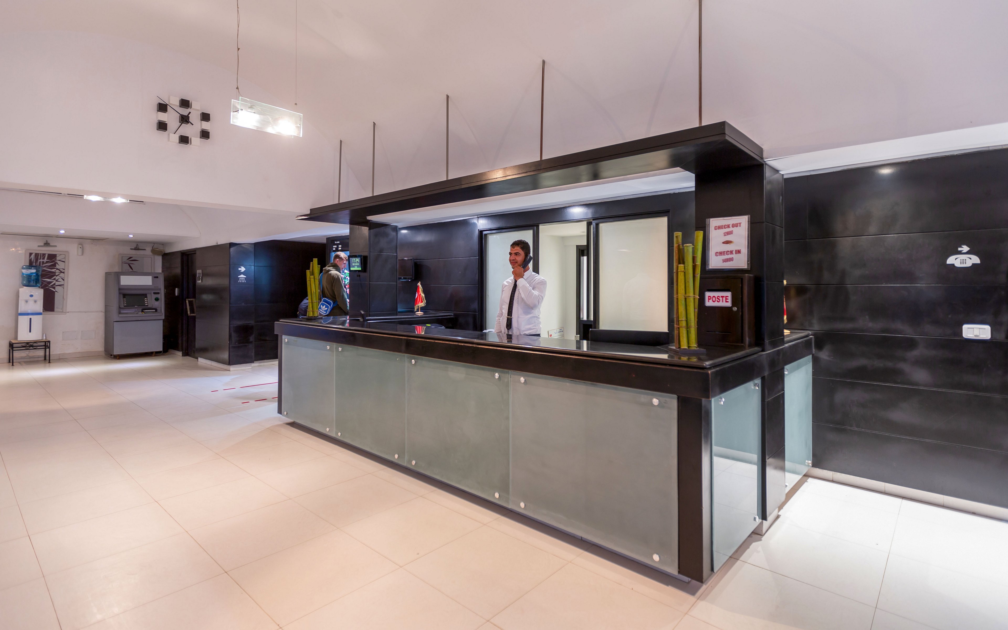 A hotel receptionist stands behind a sleek, modern front desk in a spacious lobby. The reception counter features dark panels with frosted glass and decorative tall vases. Bright overhead lighting and light-colored floor tiles create a clean, contemporary atmosphere. Various wall-mounted signs and equipment are visible in the background, suggesting a professional hospitality setting.