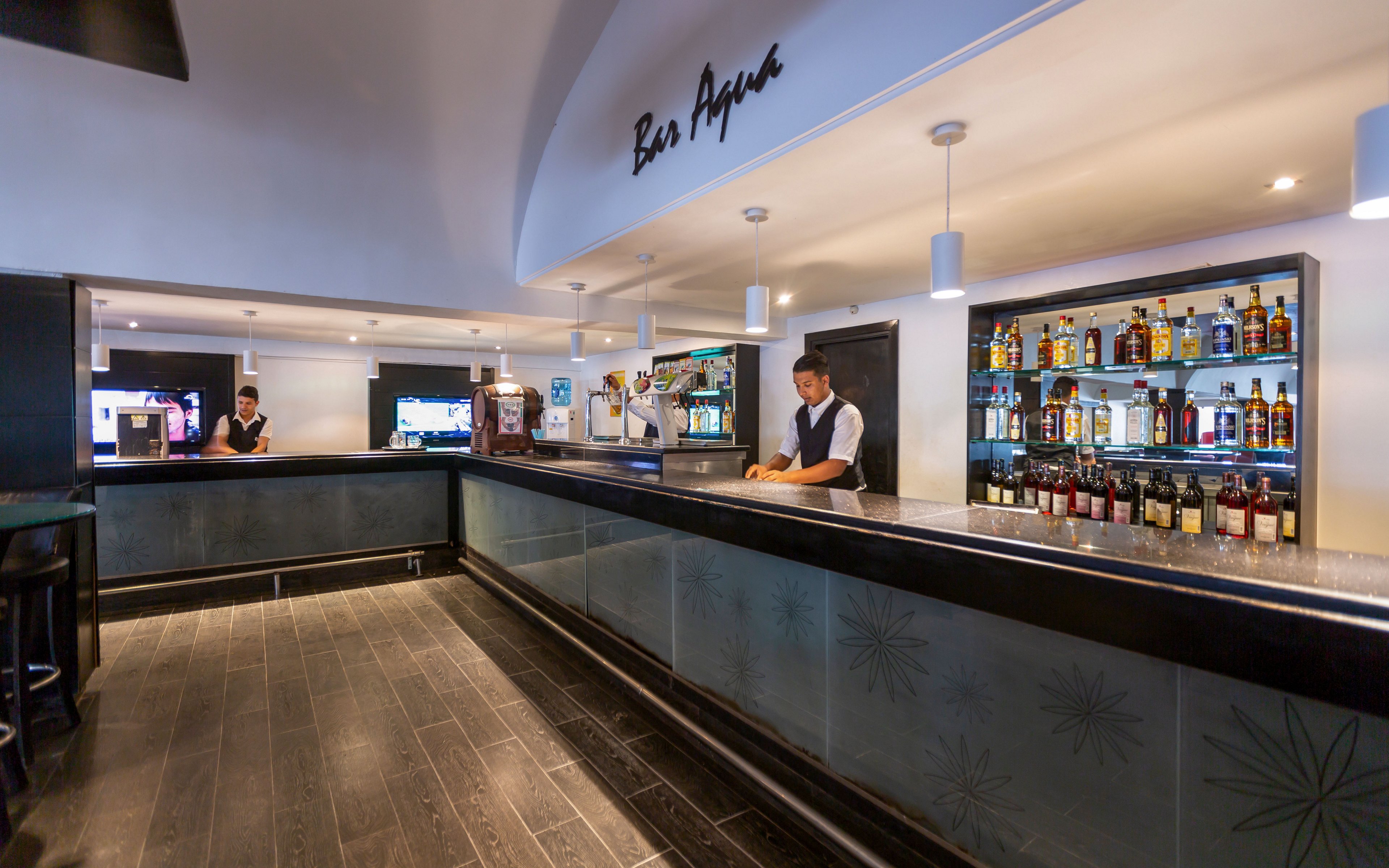 A spacious contemporary bar interior features a long counter with a bartender standing behind it. Shelves in the background display a wide variety of liquor bottles and glassware. Pendant lights hang from the ceiling, illuminating the sleek dark flooring and minimalist decor. Another staff member is visible further down the bar, contributing to the professional atmosphere. The overall scene suggests a clean, stylish venue ready to serve guests.
