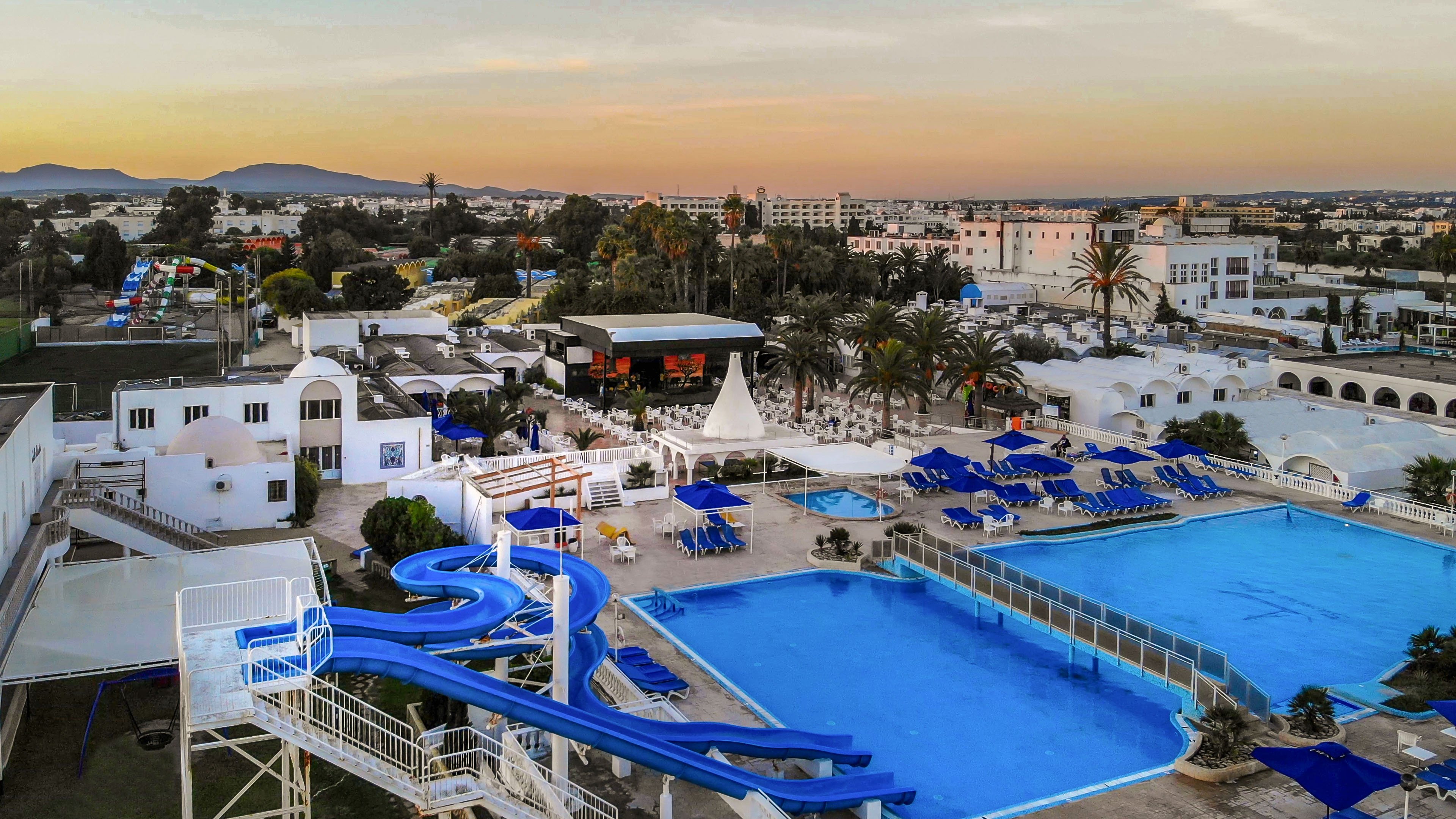 A large resort complex is shown from above at sunset, featuring expansive outdoor swimming pools and winding blue waterslides. Rows of sun loungers and umbrellas surround the pools, creating a relaxed vacation atmosphere. White low-rise buildings, palm trees, and landscaped areas extend into the background, suggesting a coastal or warm-weather destination. The sky is softly lit with warm tones, enhancing the tranquil holiday mood.