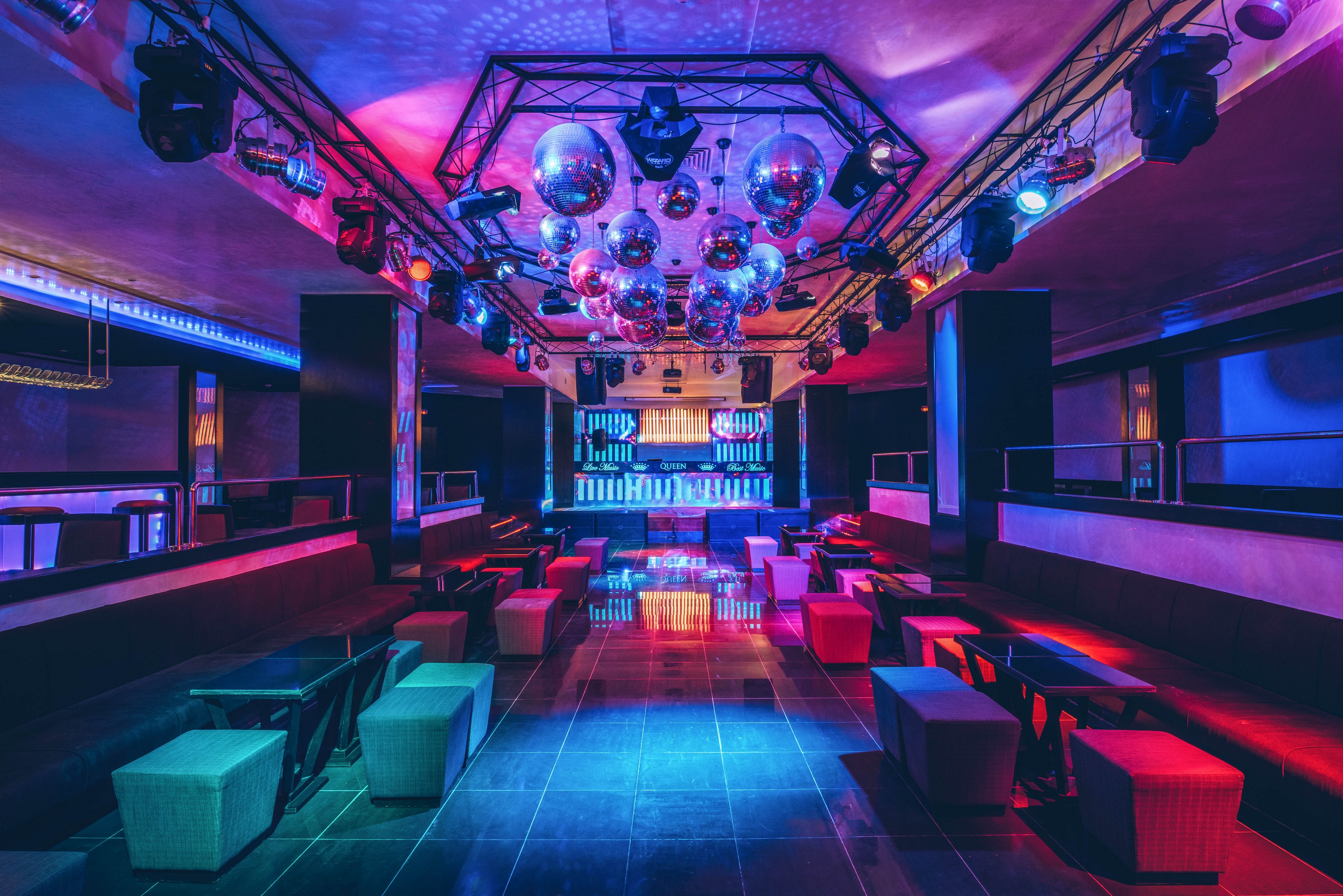 A modern nightclub interior is shown empty, with rows of low benches and tables arranged around a central open floor. The space is illuminated by vibrant blue, pink, and purple lighting that reflects off glossy tiles. Numerous disco balls and spotlights hang from the ceiling, creating a dynamic party atmosphere. At the far end, a lit bar or DJ stage area glows with vertical light panels, emphasizing the club’s contemporary design.
