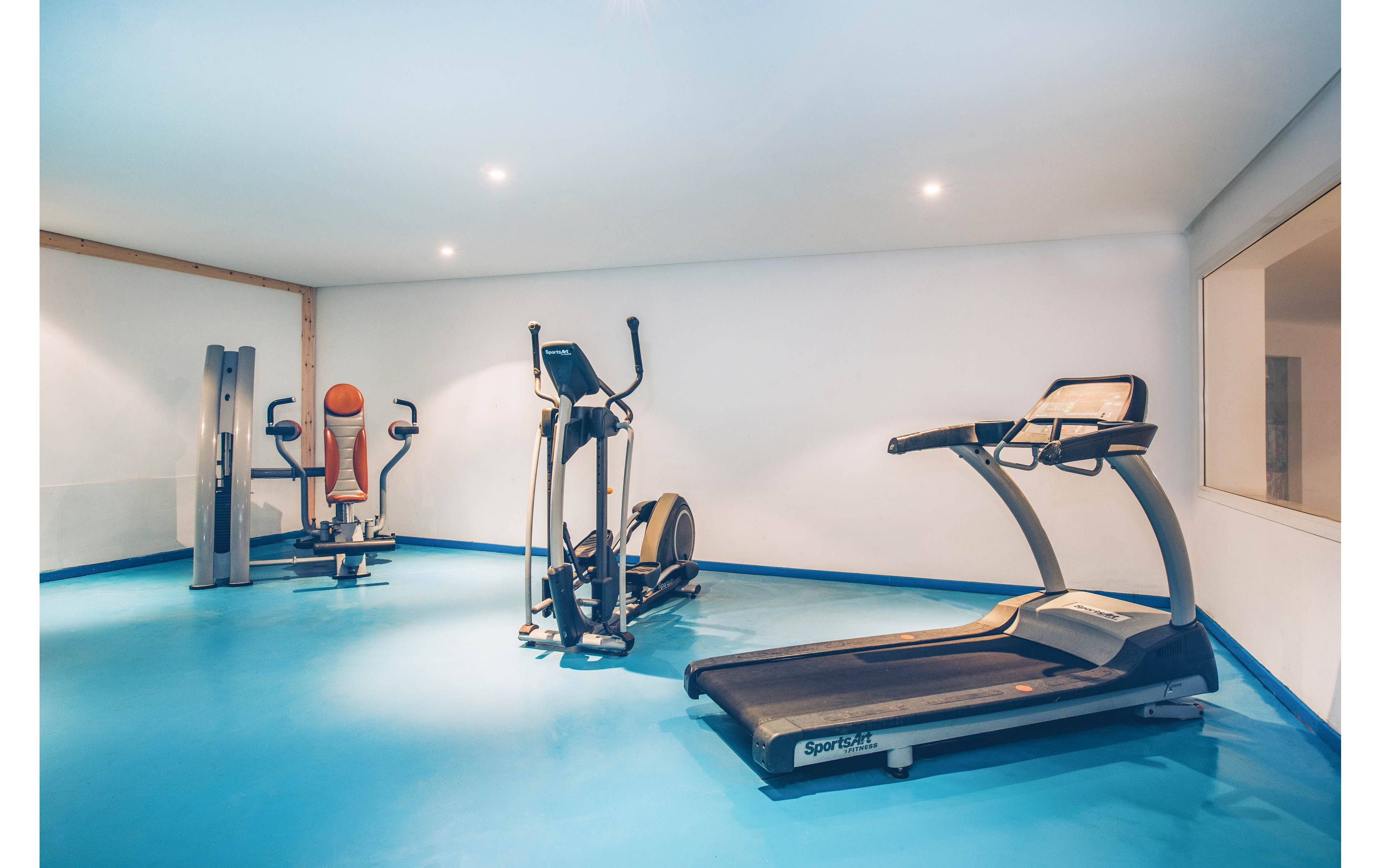 A clean, modern gym room features several pieces of cardio and strength equipment on a bright blue floor. A treadmill, elliptical trainer, and multi-station weight machine are arranged against white walls under recessed ceiling lights. The space appears minimalistic and uncluttered, with no people present. A large interior window on the right side adds openness to the workout area.