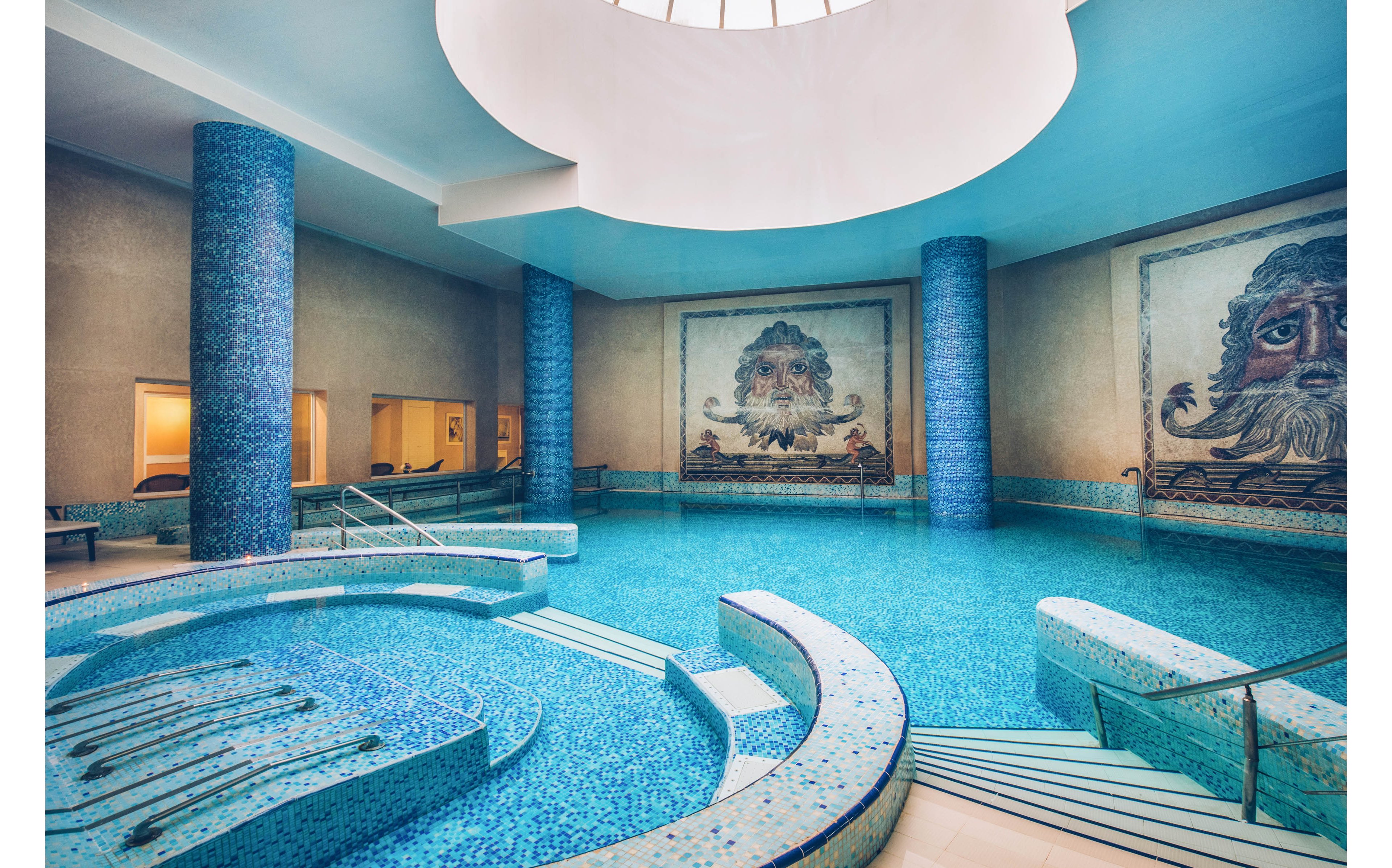 Luxurious indoor spa pool featuring curved seating and steps is shown under a large circular skylight. The turquoise-tiled water area is surrounded by tall blue mosaic columns and smooth beige walls. Large wall mosaics depicting stylized faces add an artistic focal point to the serene space. Warm ambient lighting from adjacent rooms enhances the relaxing atmosphere.